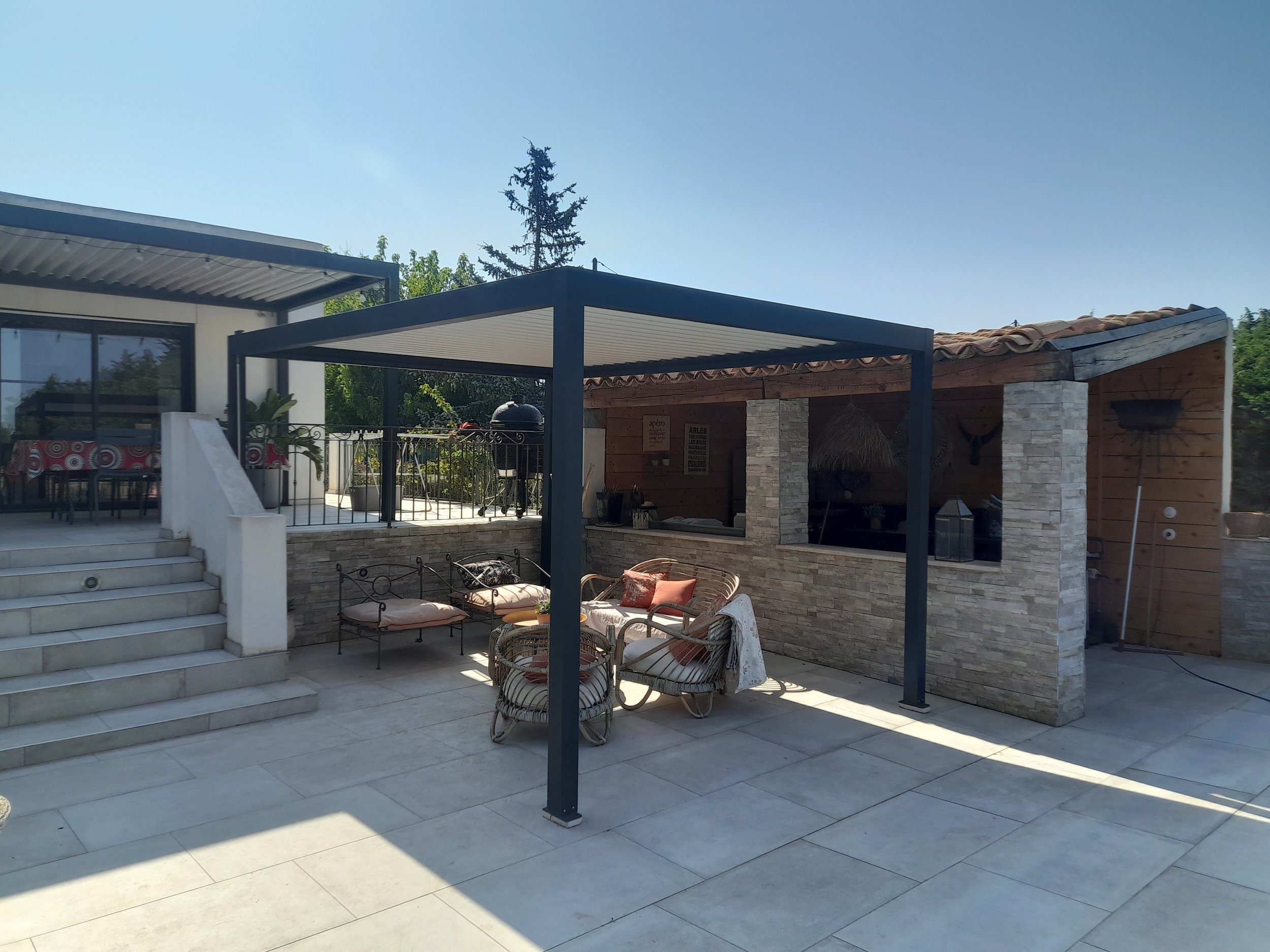 Une terrasse extérieure avec mobilier en rotin, un canapé et des chaises, sous une structure en métal avec pergola. En arrière-plan, un mur en pierre et des plantes. Le ciel est clair et ensoleillé.