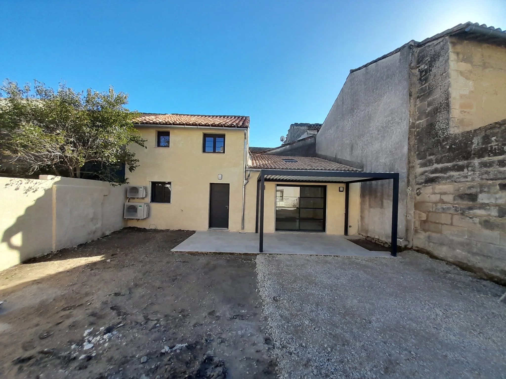 Vue de la cour extérieure d'une maison avec sol non pavé, un mur en pierre, une maison jaune avec fenêtres et toiture en tuiles, un arbre et un ciel clair.