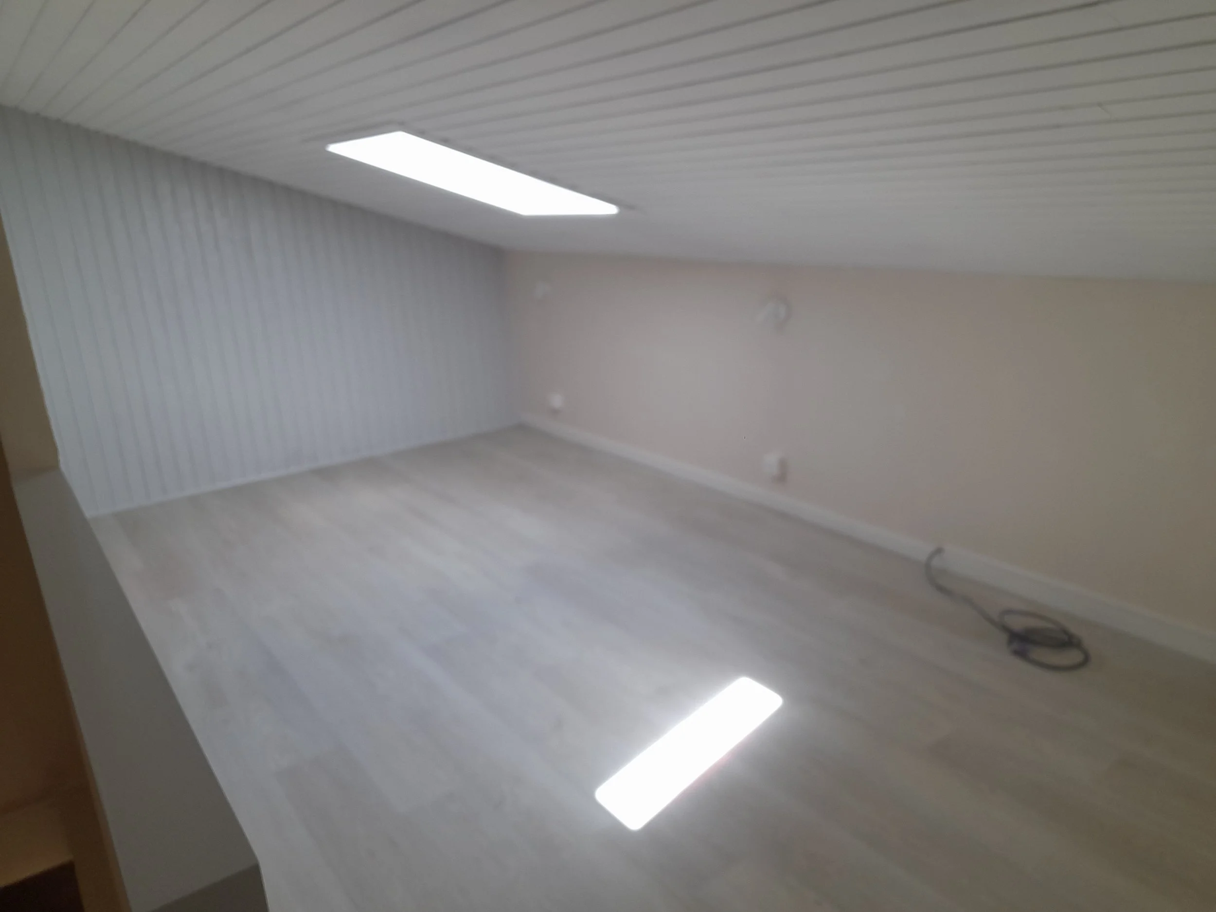 Une pièce probablement un bureau ou un espace de travail vide avec un plafond en lambris blanc, un éclairage fluorescent, un mur blanc clair, et des câbles électriques au sol.