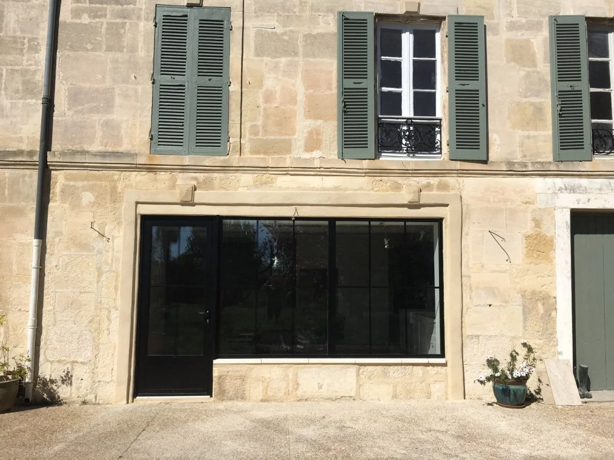 Façade d'une maison en pierre avec des volets verts, fenêtres à doubles volets et une grande fenêtre noire au sol.