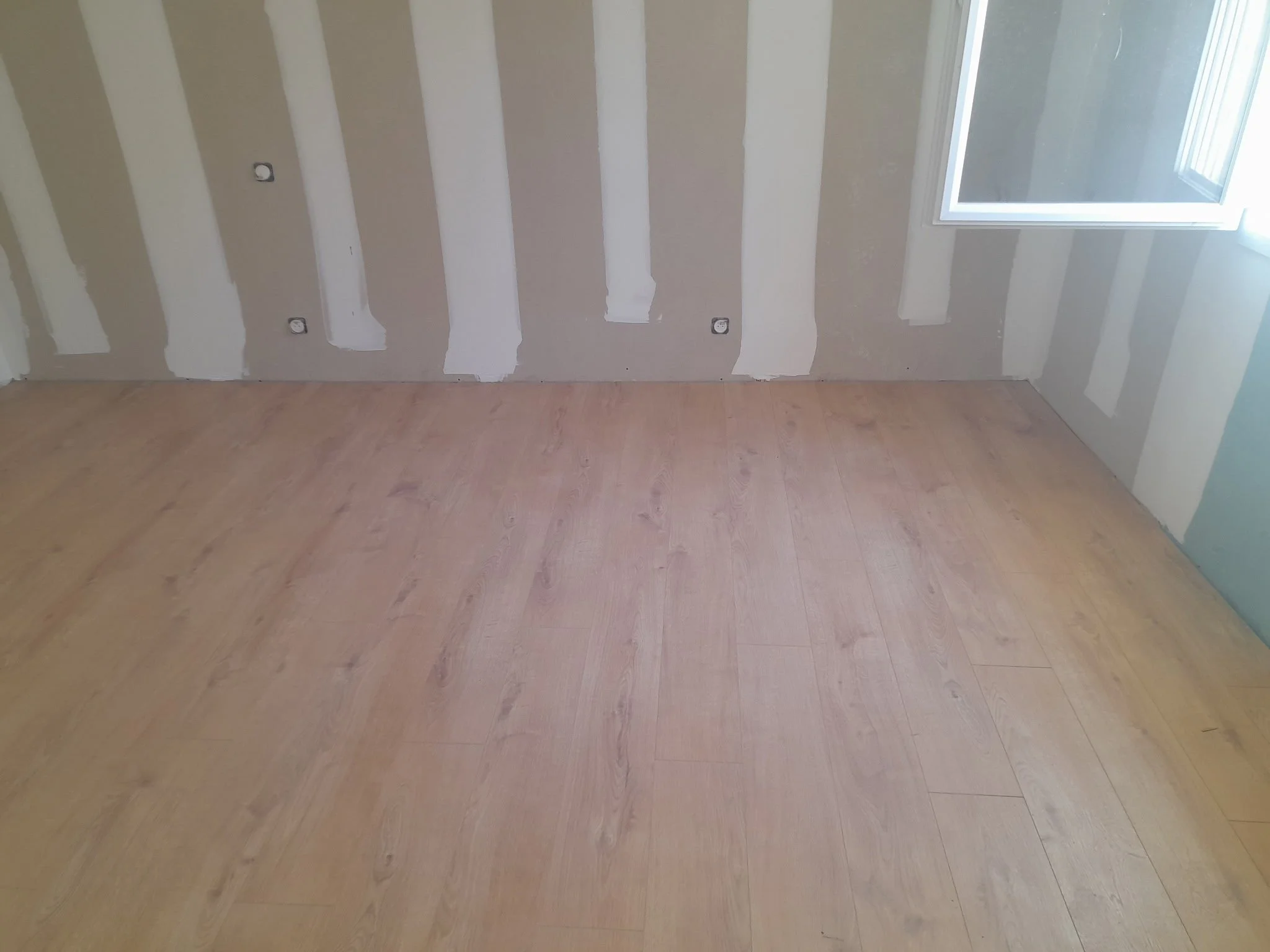 Une pièce avec un sol en bois clair, des murs en plâtre partiellement peints en blanc et beige, une grande fenêtre laissant entrer la lumière naturelle.