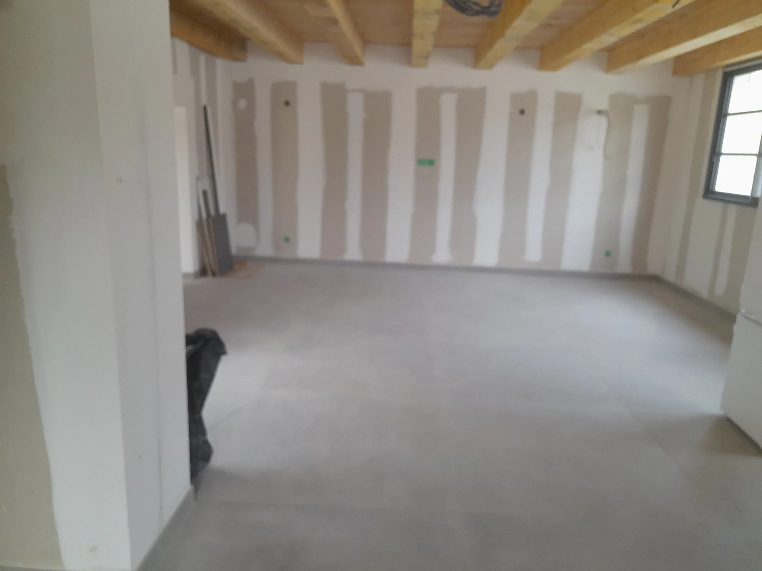 Une pièce en cours de construction ou de rénovation, avec des murs en plaques de plâtre non peintes, un plafond en bois avec des poutres apparentes, et une fenêtre laissant entrer de la lumière. On aperçoit aussi des outils ou matériaux au sol.