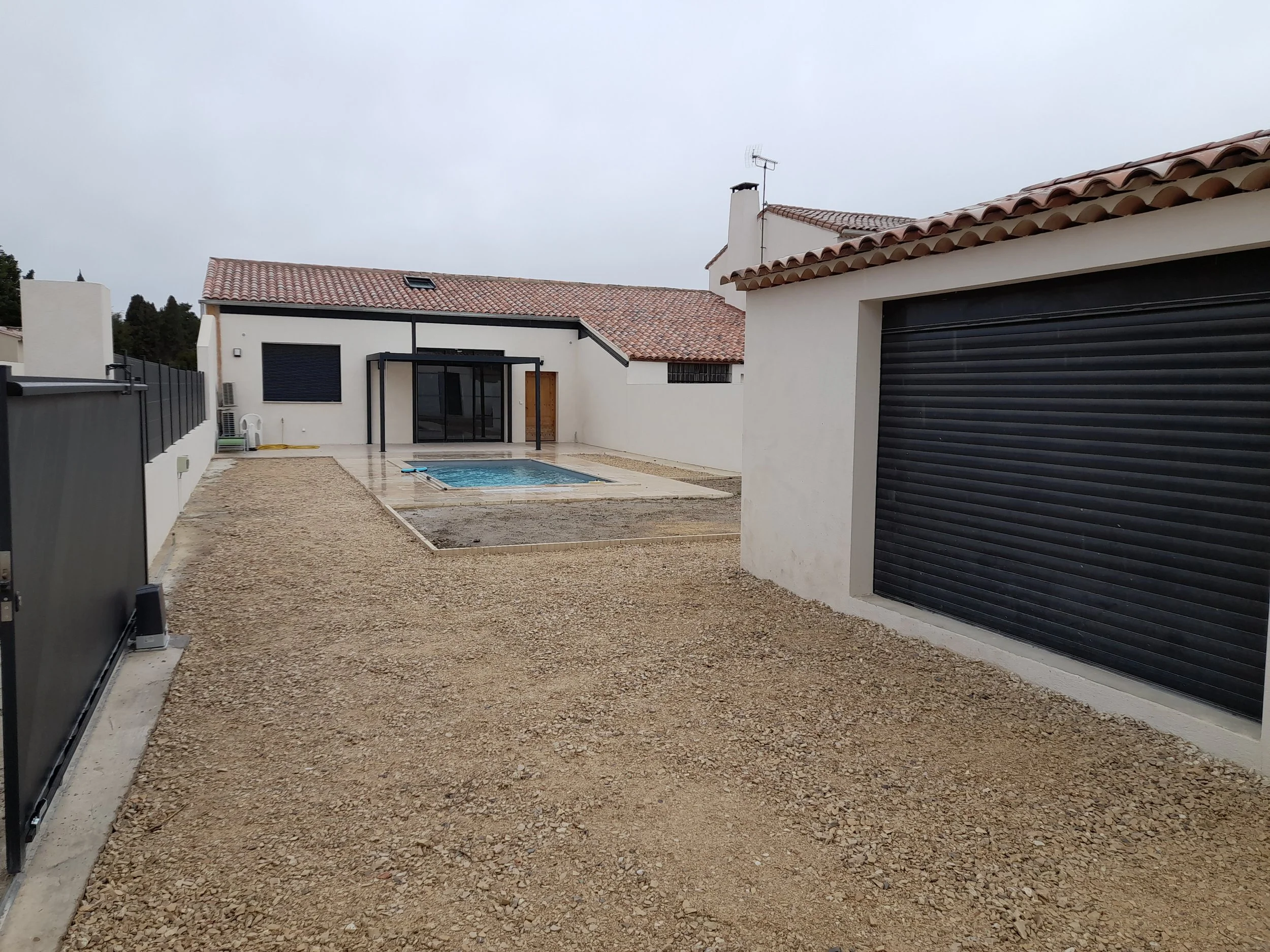 Yard with a small swimming pool, modern white house with black shutters, gravel pathway, and closed garage door, under a cloudy sky.