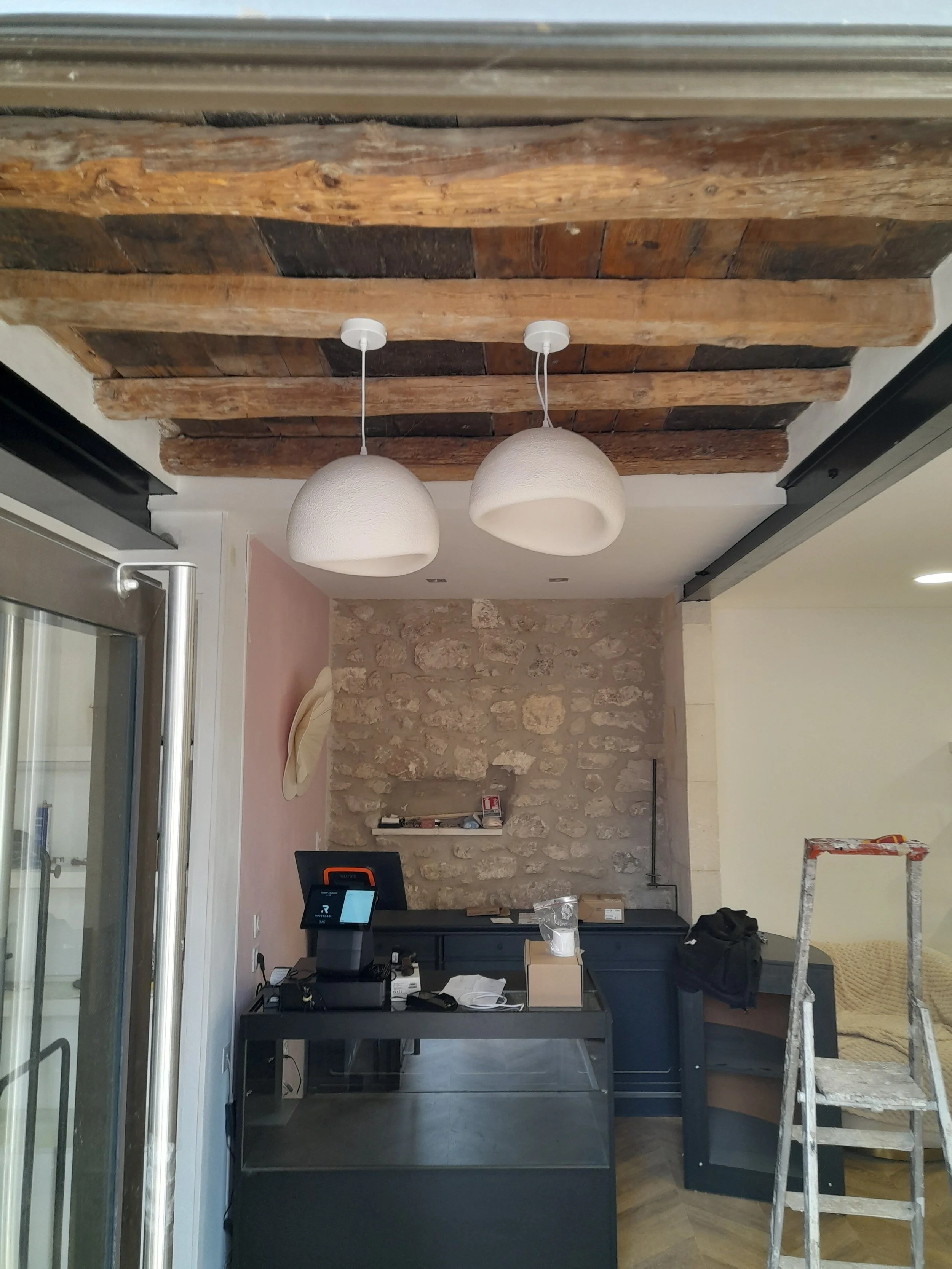 Intérieur d'un espace de travail avec un mur en pierre, un comptoir noir, deux luminaires suspendus au plafond en bois ancien, un rebord de fenêtre en verre à gauche, une échelle en métal à droite, et divers objets sur le comptoir.