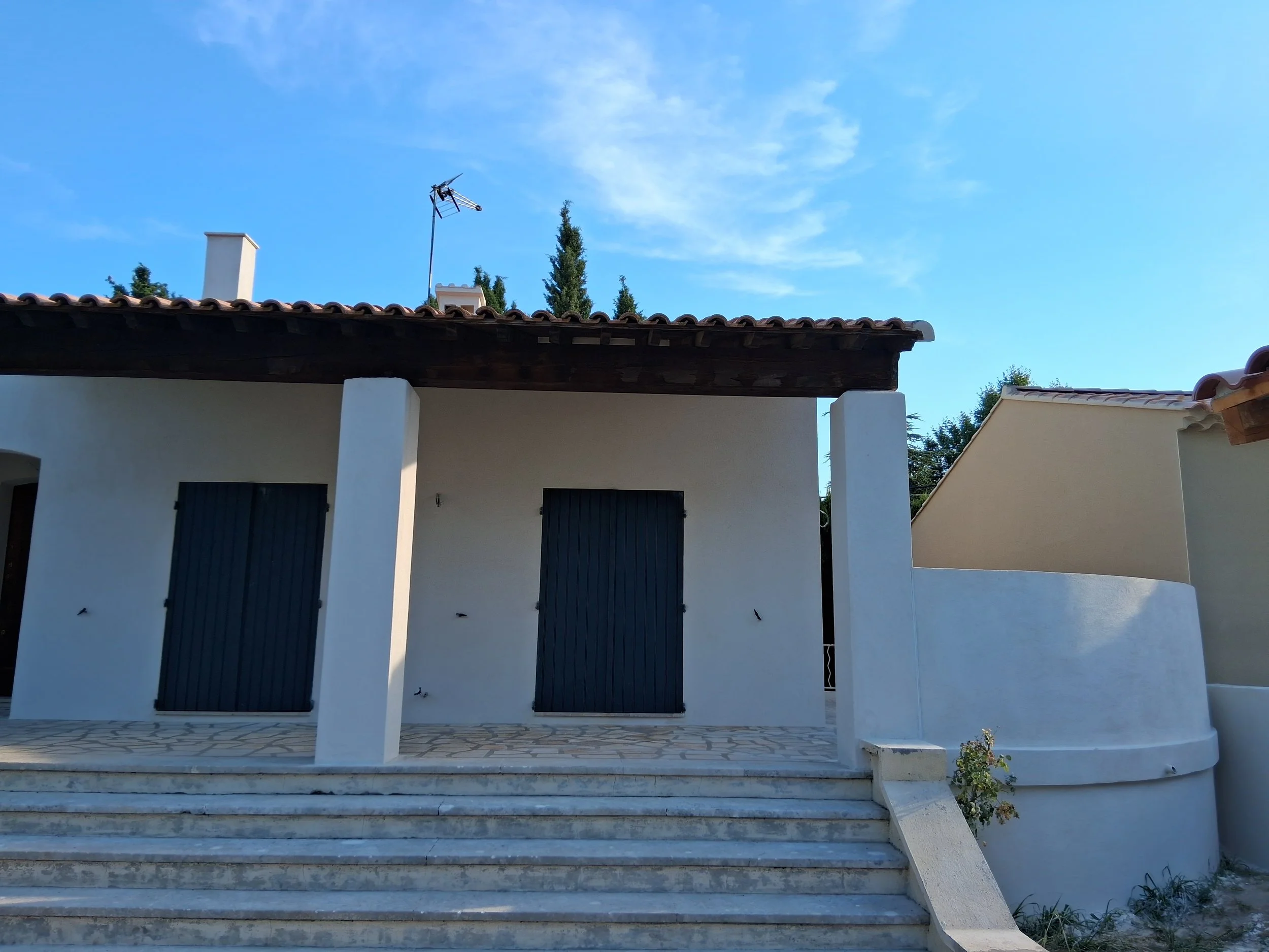 Une maison moderne avec une façade blanche, des volets noirs et un toit en tuiles, avec un escalier en pierre menant à la porte d'entrée et un ciel bleu clair avec quelques nuages.