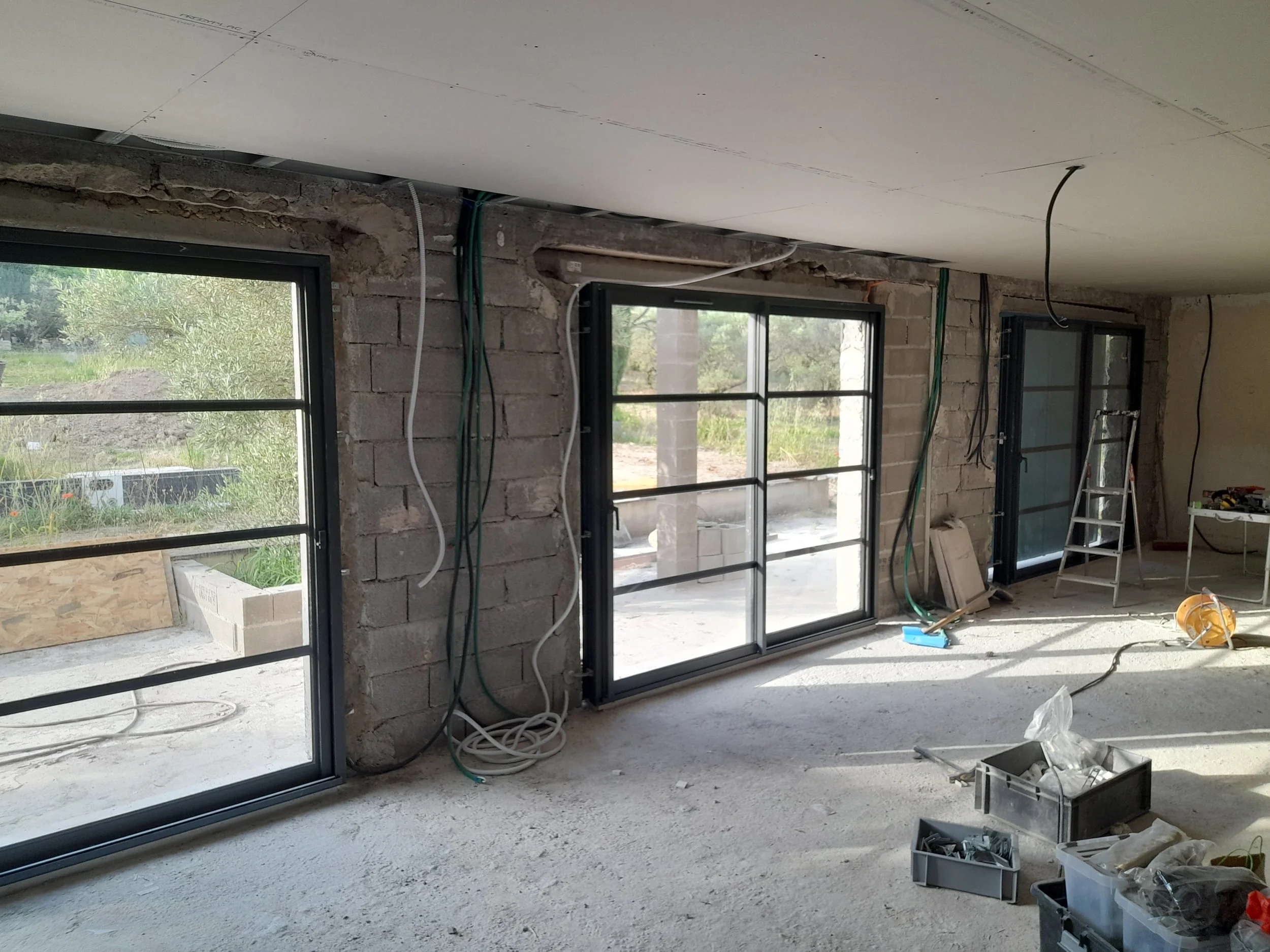 Une pièce en construction avec fenêtres, des câbles suspendus au plafond, des murs en briques, et des outils de construction.