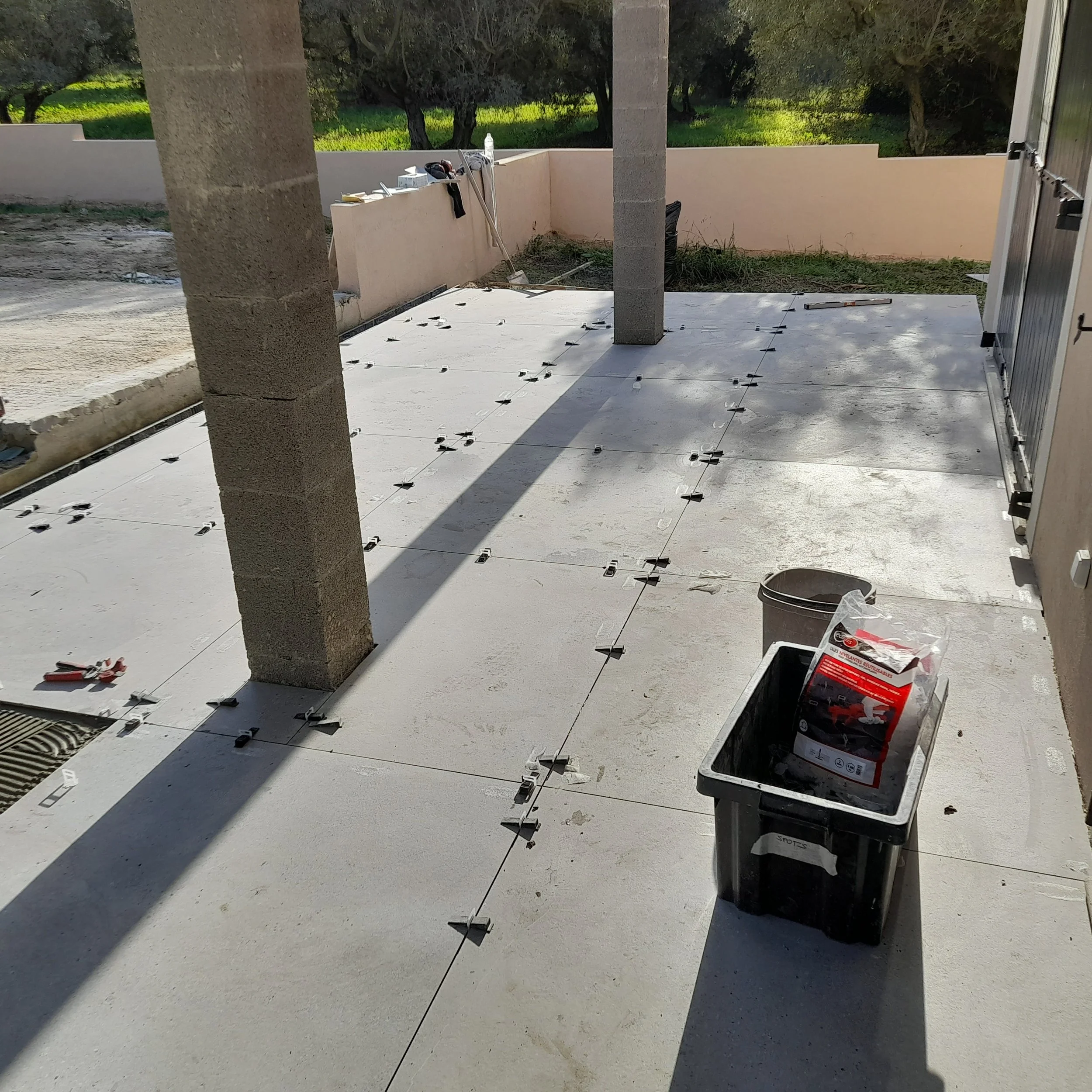 Une terrasse en cours de construction avec des rails pour la pose de carreaux de sol, outils et matériaux de construction, entourée d'un mur beige et de deux piliers en béton, avec un peu de végétation en arrière-plan.