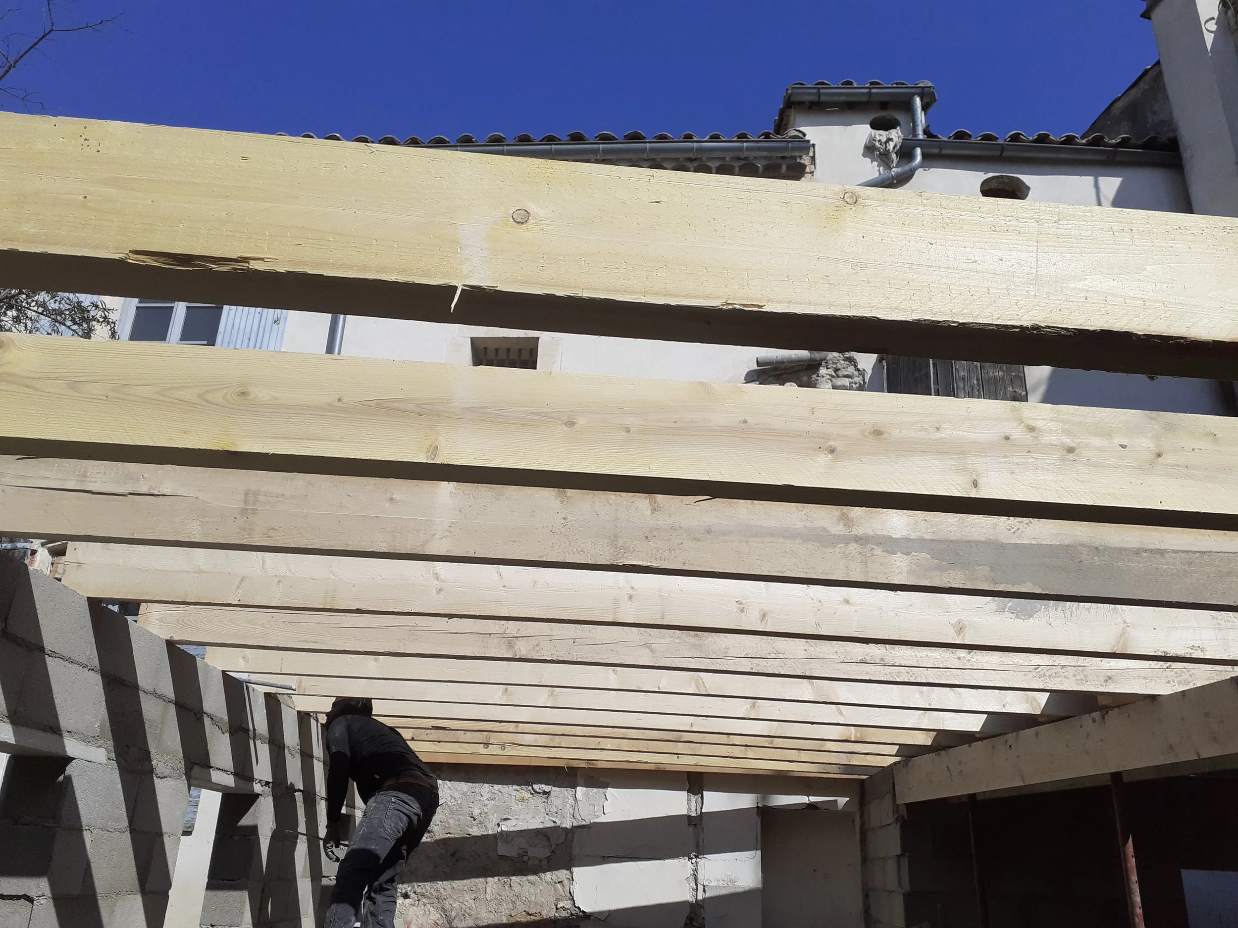 Construction de clôture avec des planches en bois clouées verticalement, le ciel bleu en arrière-plan, et une personne travaillant sur le chantier.