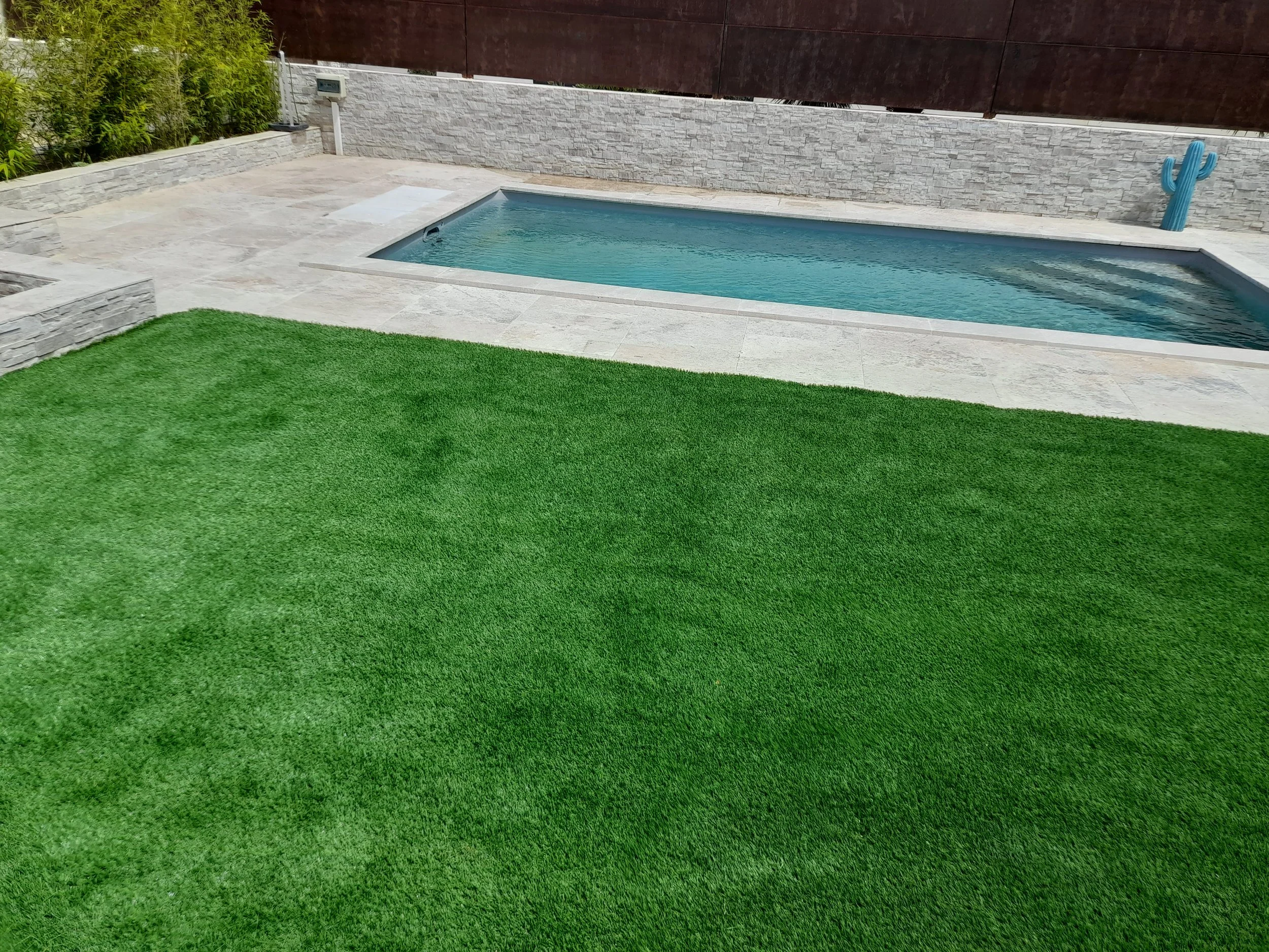 Petite piscine dans un jardin avec une pelouse verte, un mur en pierre blanc et une clôture en bois foncé.