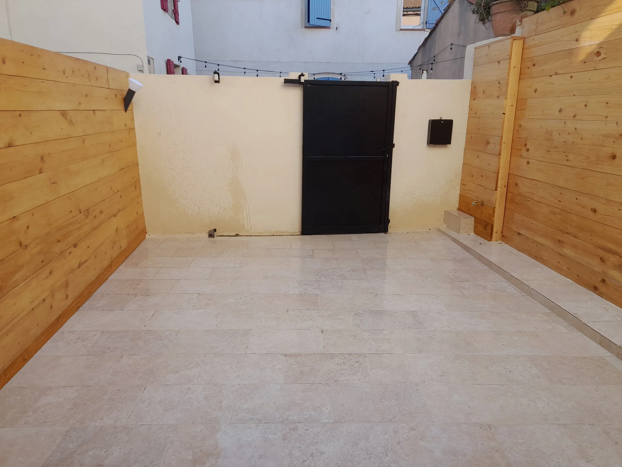 Un petit espace extérieur avec un mur blanc, un portail en bois noir, des murs en bois et une chaîne lumineuse suspendue au-dessus.