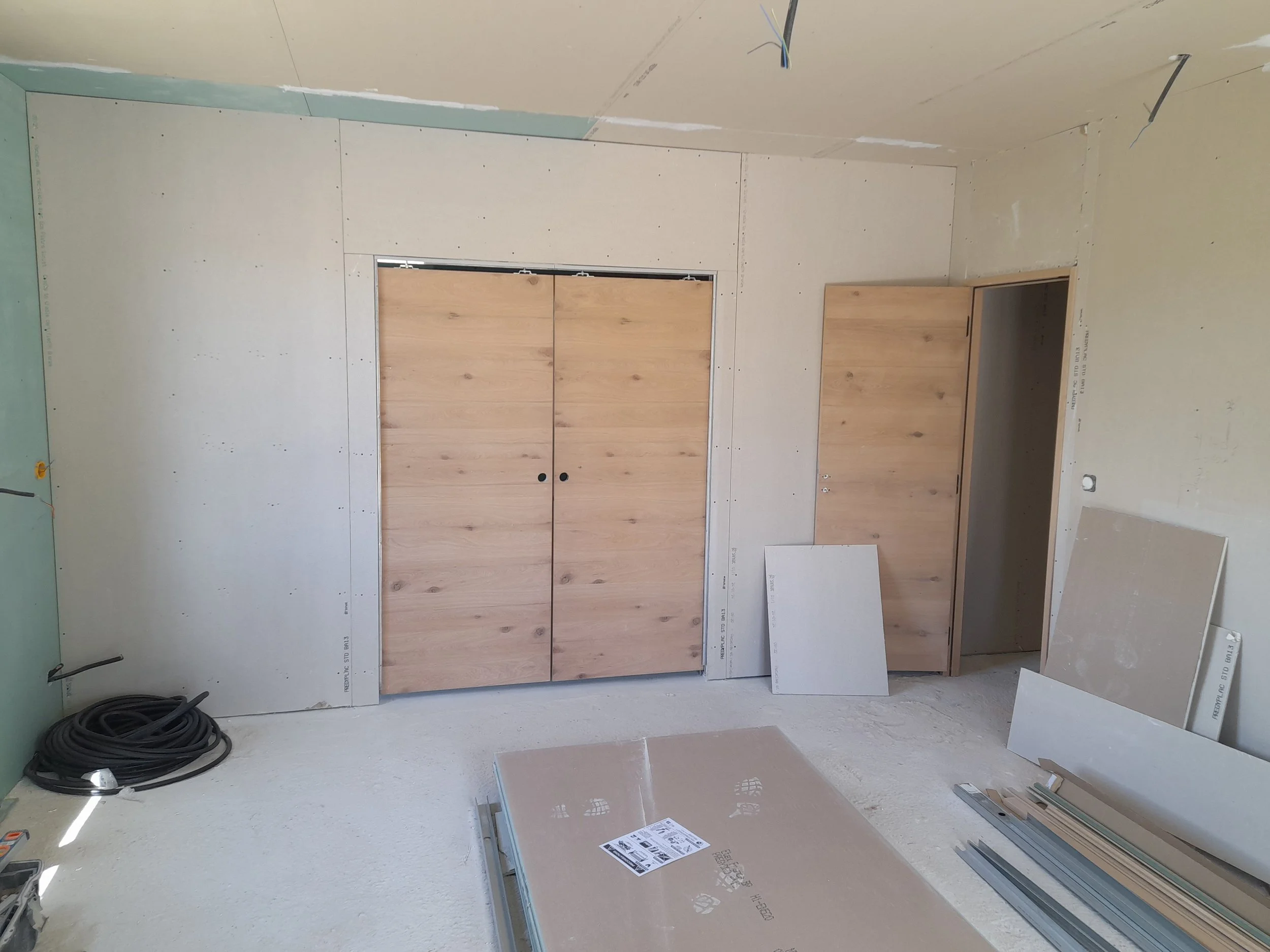 Une pièce en cours de construction avec des murs en plaques de plâtre, des panneaux de bois en préparation pour une porte ou un placard, des câbles électriques suspendus au plafond, et des outils de construction sur le sol.