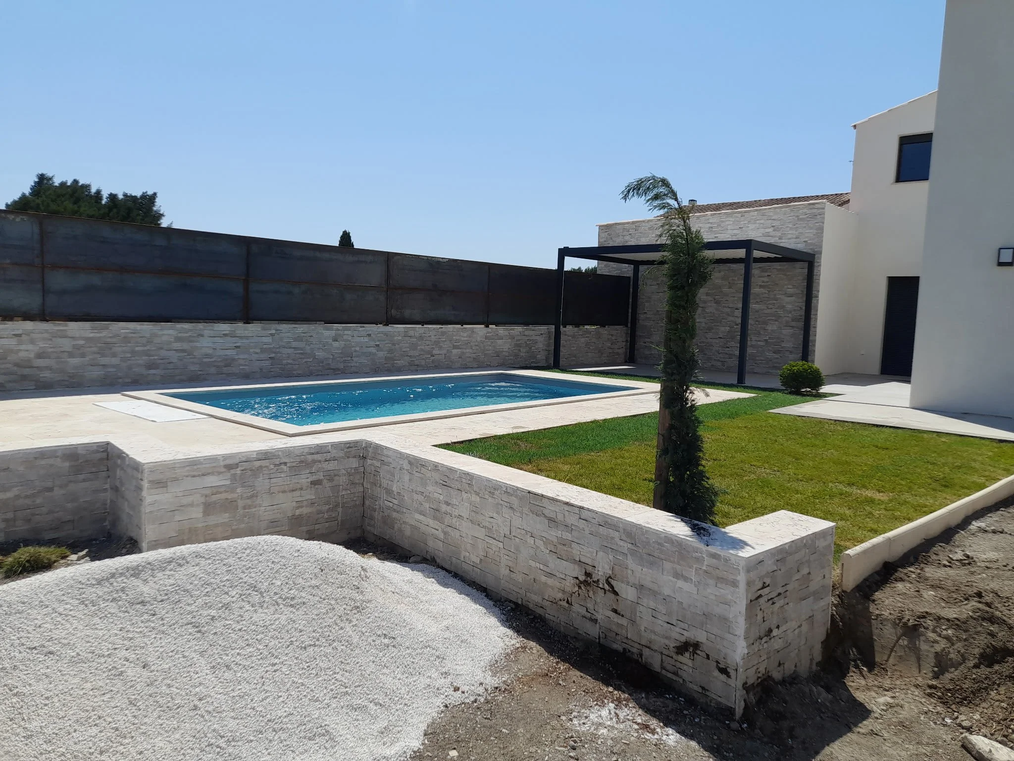 Une maison moderne avec une piscine, un mur en pierre, une pergola noire, et un arbre jeune dans le jardin.