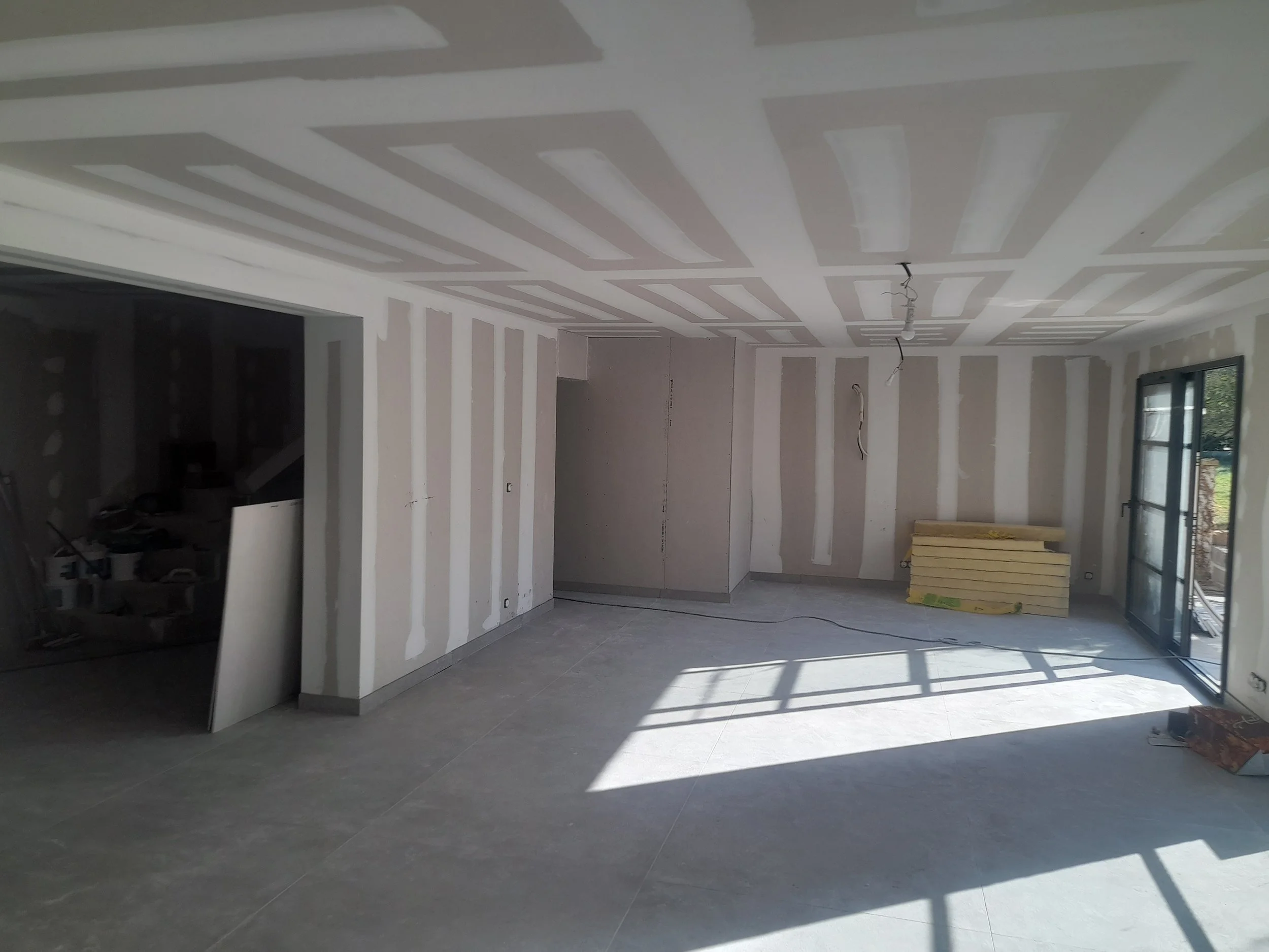 Intérieur d'une pièce en cours de construction avec murs en plaques de plâtre, plafond en plaques de plâtre avec joints, et une fenêtre laissant passer la lumière naturelle.