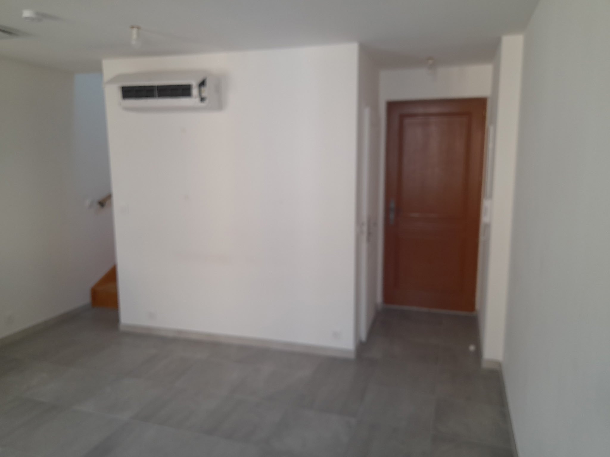 Intérieur d'un appartement avec murs blancs, plancher en carrelage gris, porte en bois, et un appareil de climatisation mural.