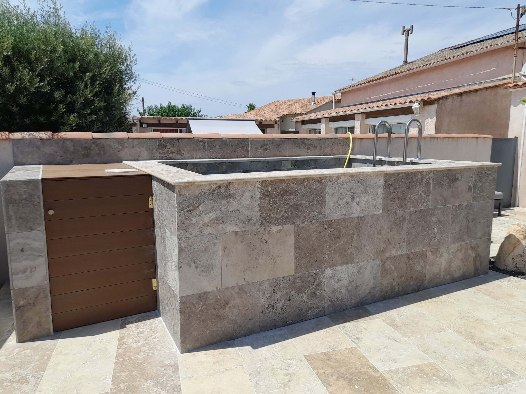 Un espace extérieur avec un jacuzzi construit en pierres, une terrasse en carrelage, et des maisons avec des toits en tuiles en arrière-plan, sous un ciel partiellement nuageux.