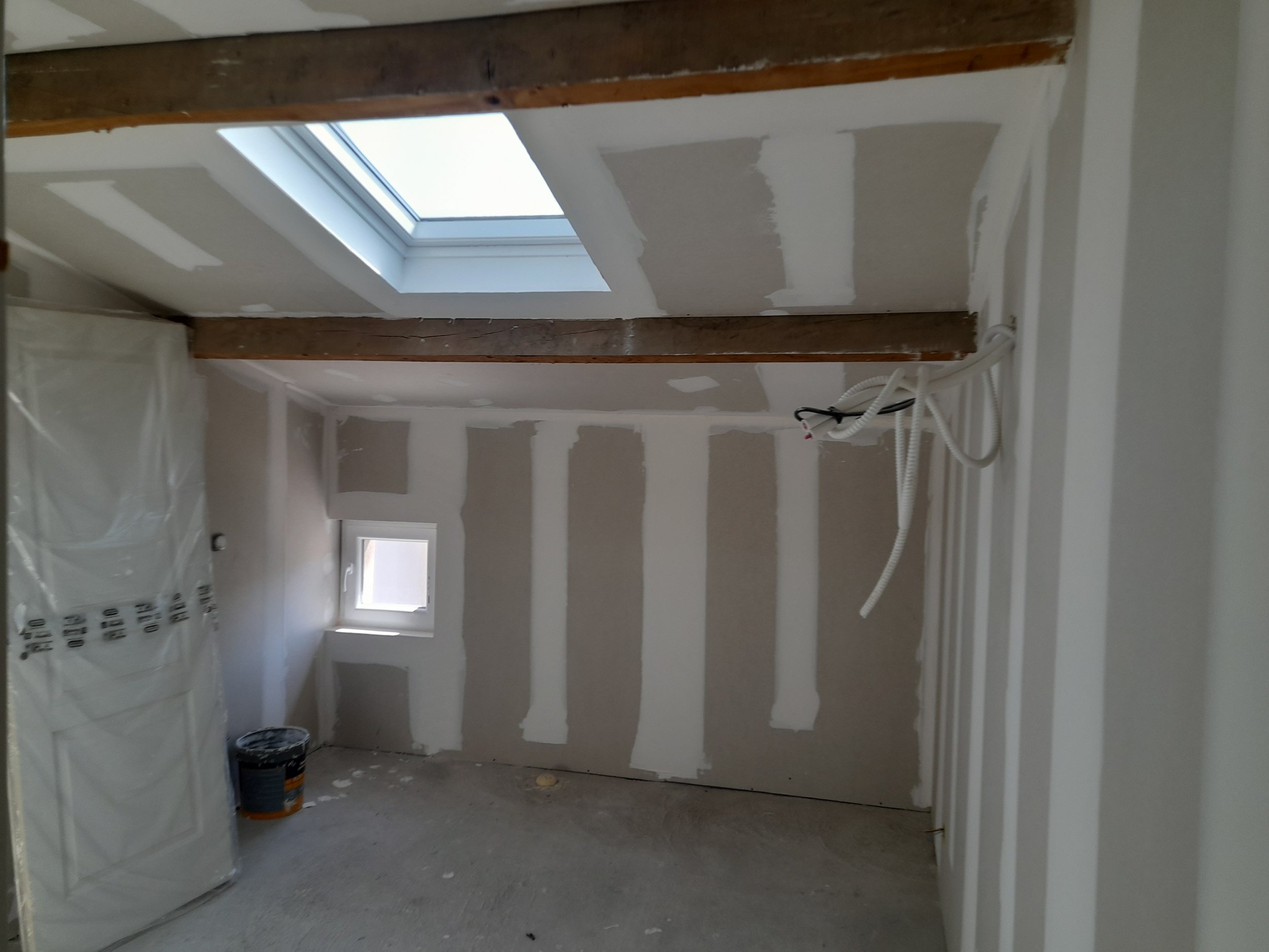 Une pièce en cours de rénovation avec des murs en placoplâtre, des poutres apparentes, une fenêtre de toit, des câbles électriques suspendus et des pots de peinture au sol.