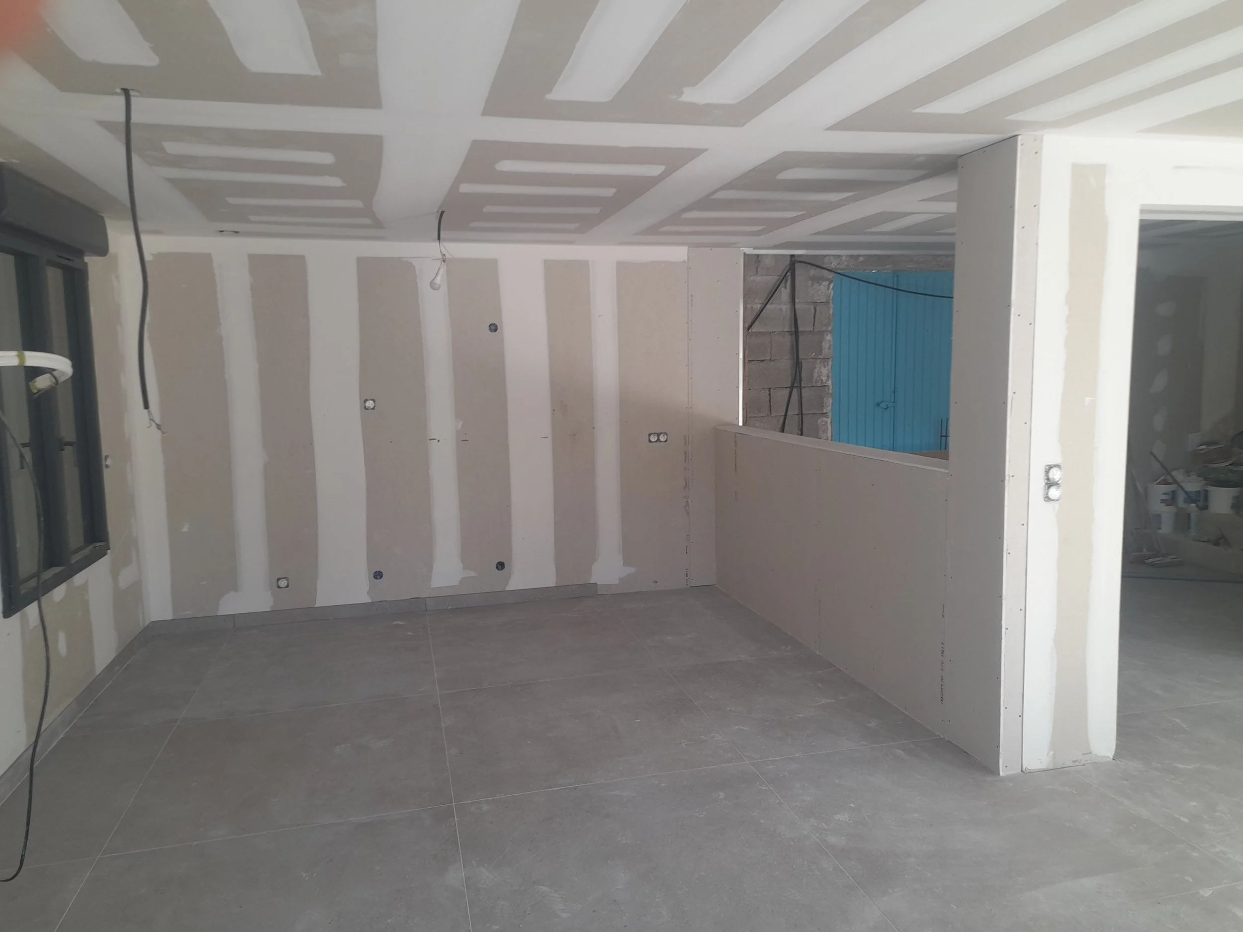 Intérieur d'une pièce en construction avec murs en plâtre, plafond suspendu en cours d'installation, et carrelage gris au sol.