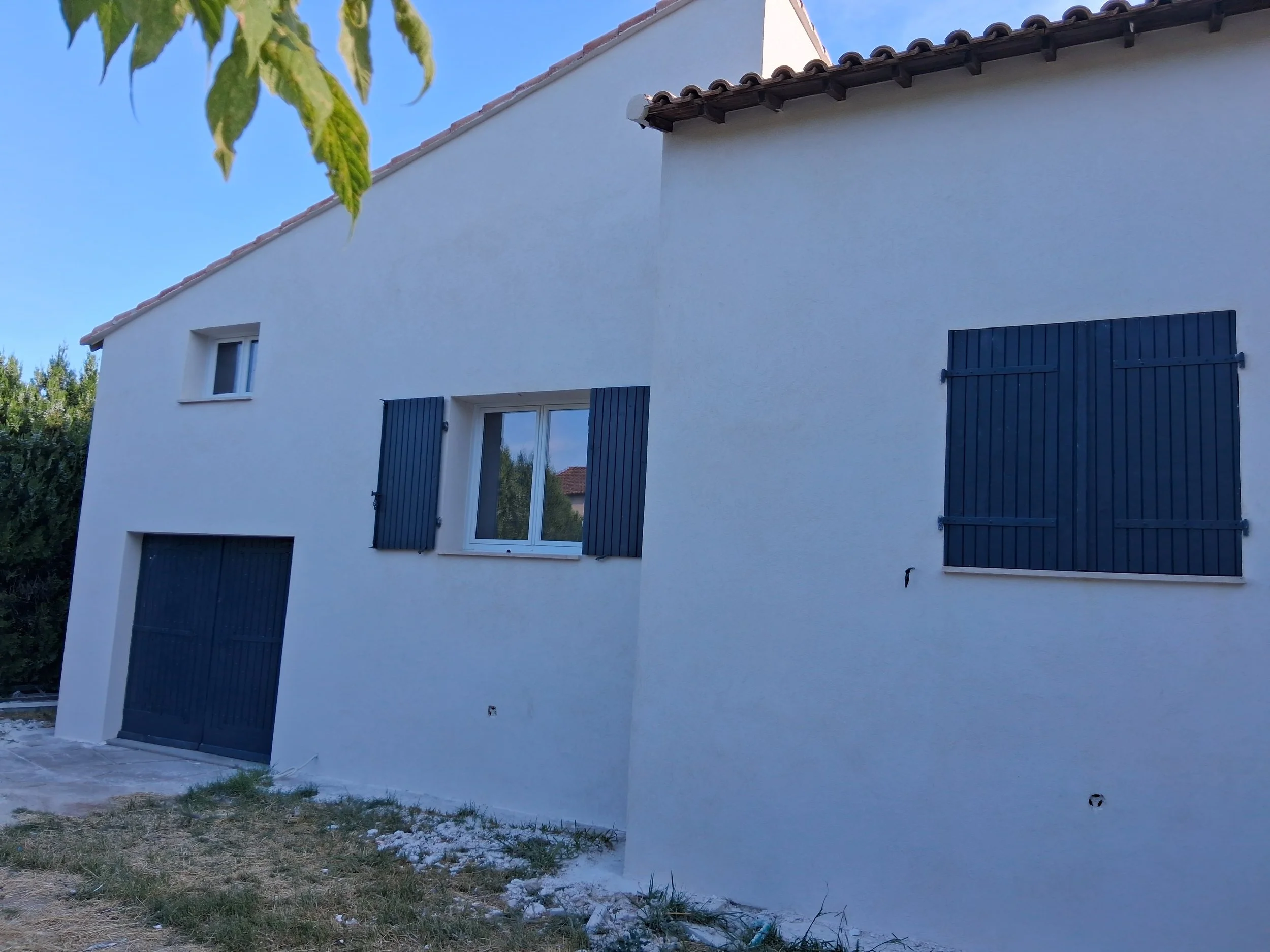 Vue d'une maison moderne avec murs blancs, fenêtres avec volets bleus, toiture en tuiles et un petit jardin avec de l'herbe et des pierres.