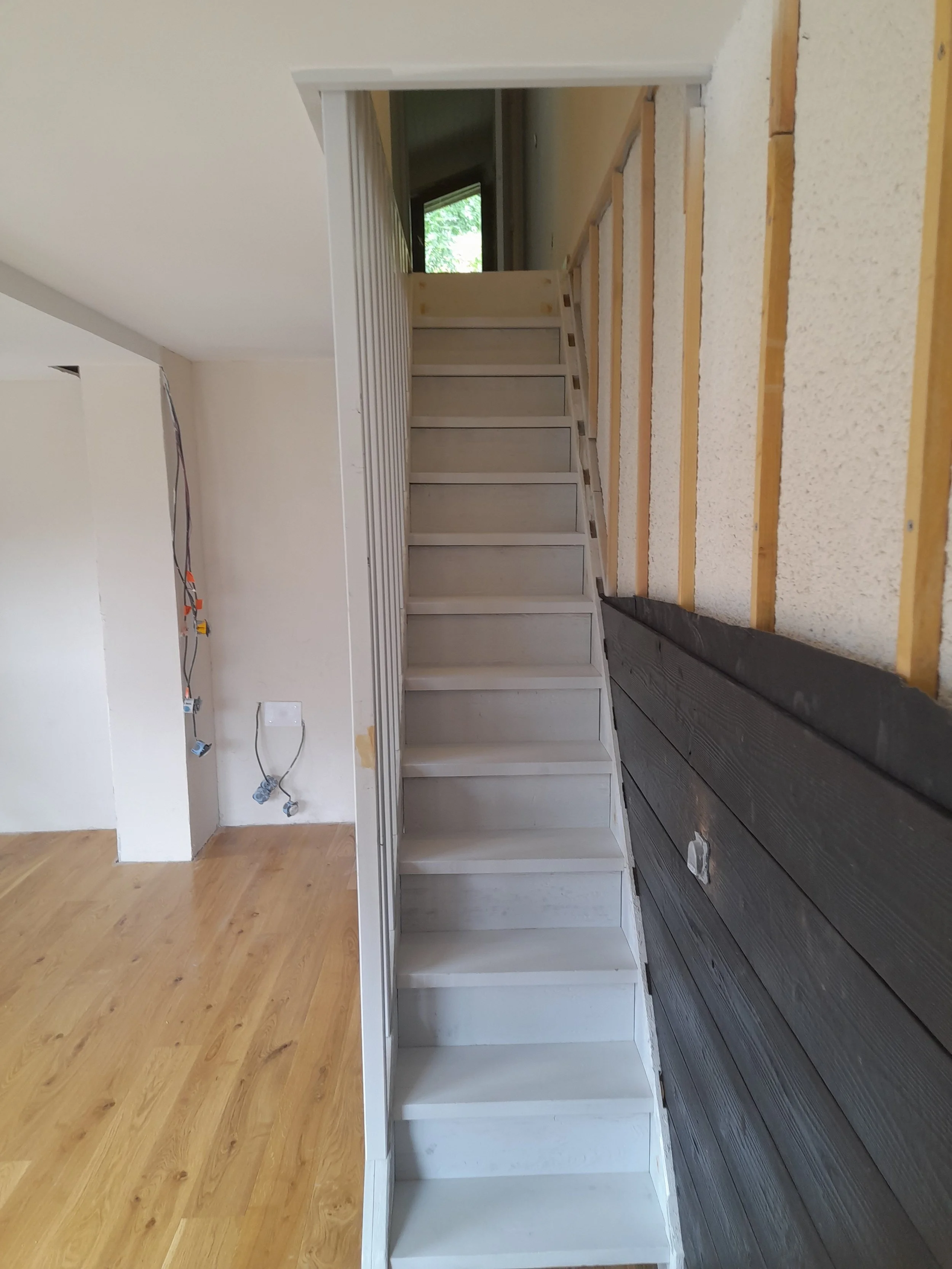 Étage d'une bibliothèque en construction avec des étagères en bois non peintes et un sol en carrelage noir, dans une pièce inachevée avec des murs beige et une fenêtre offrant une lumière naturelle.