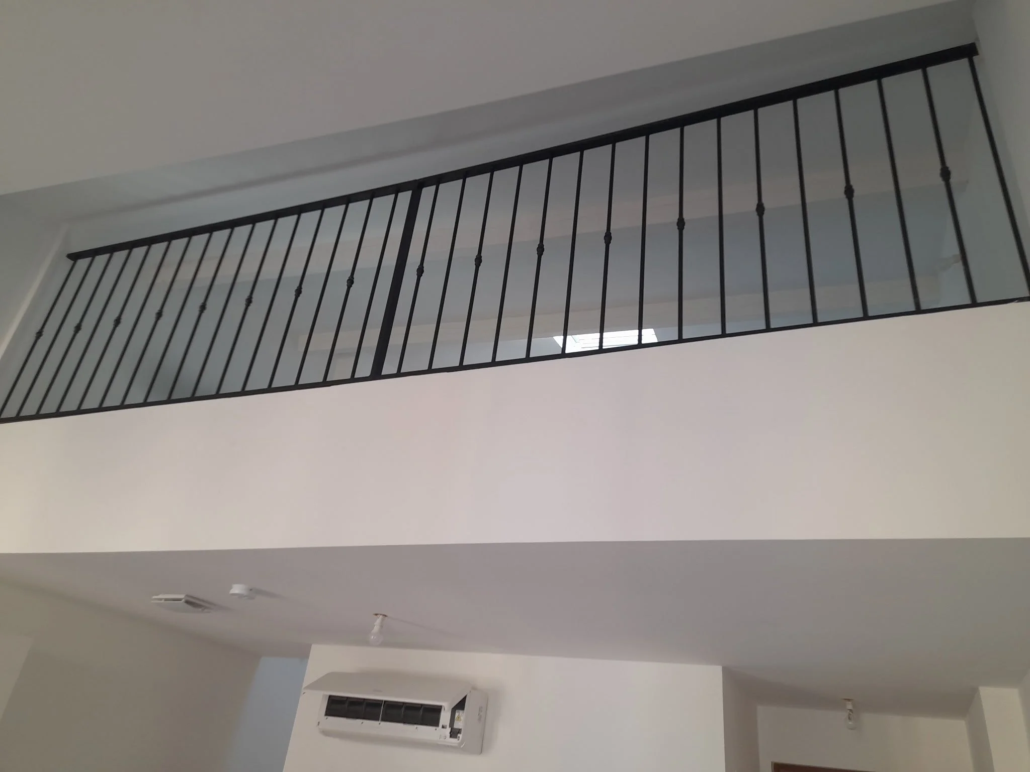 Vue du plafond d'une pièce avec une barrière en métal noir, un climatiseur mural blanc, un détecteur de fumée et un ventilateur apparent.
