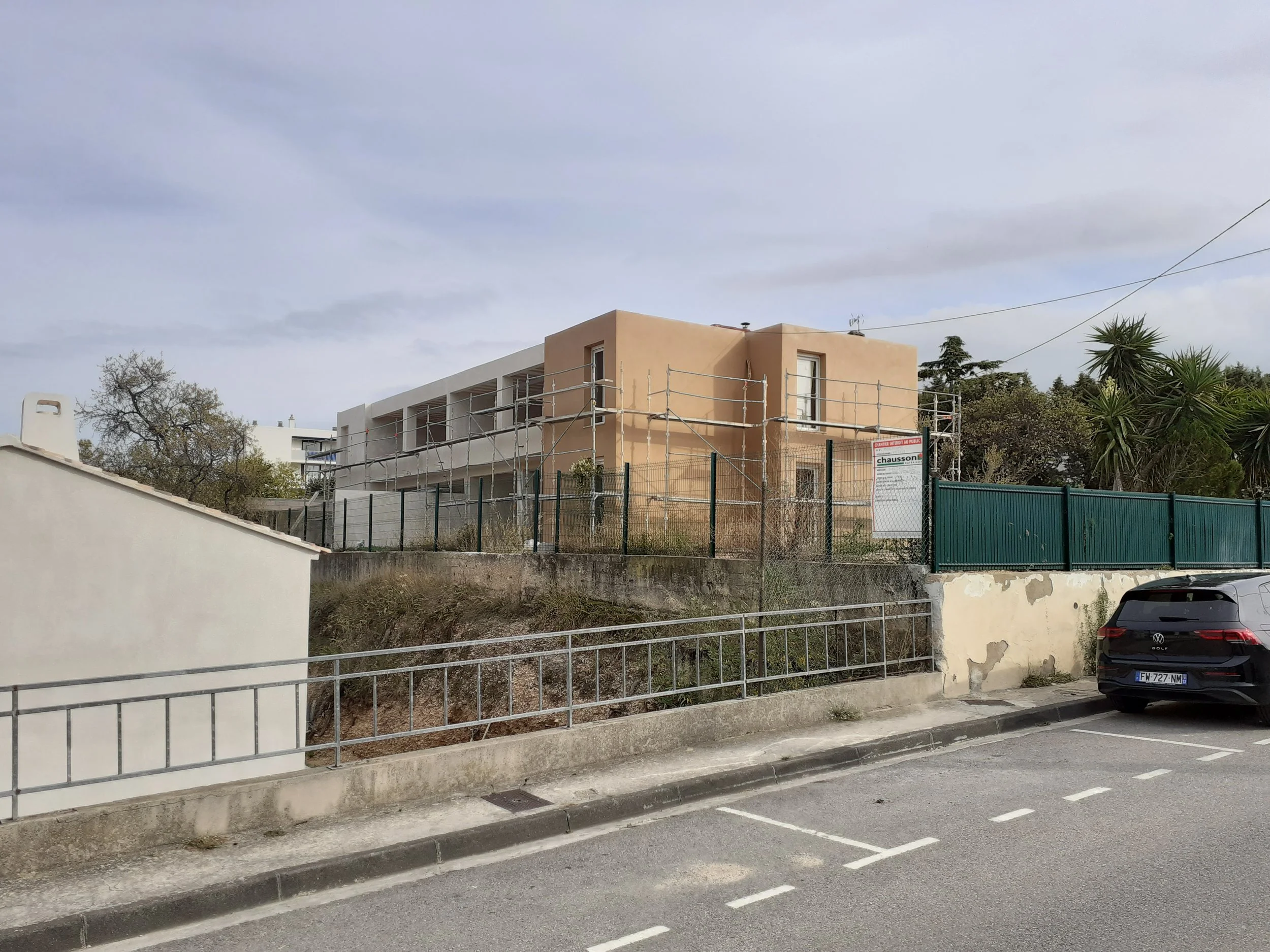 Vue d'une construction de bâtiment avec échafaudages, entourée d'un grillage, devant un mur blanc, avec quelques arbres et un parking en premier plan sous un ciel nuageux.