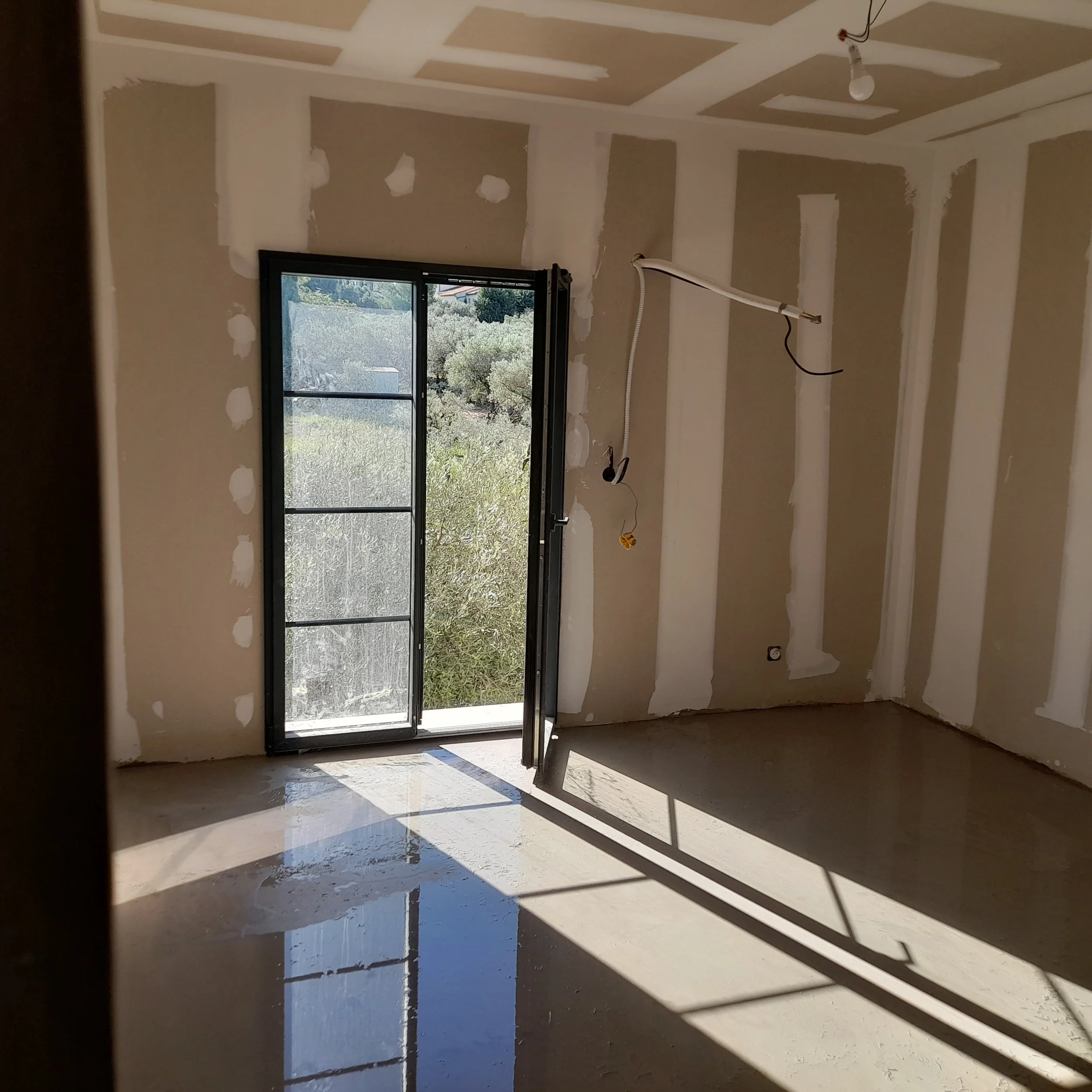 Une pièce en cours de rénovation avec un mur partiellement tapé, un plafond avec des joints en plâtre, une fenêtre donnant sur un extérieur avec des arbres, une ampoule suspendue au plafond, et des câbles électriques visibles.