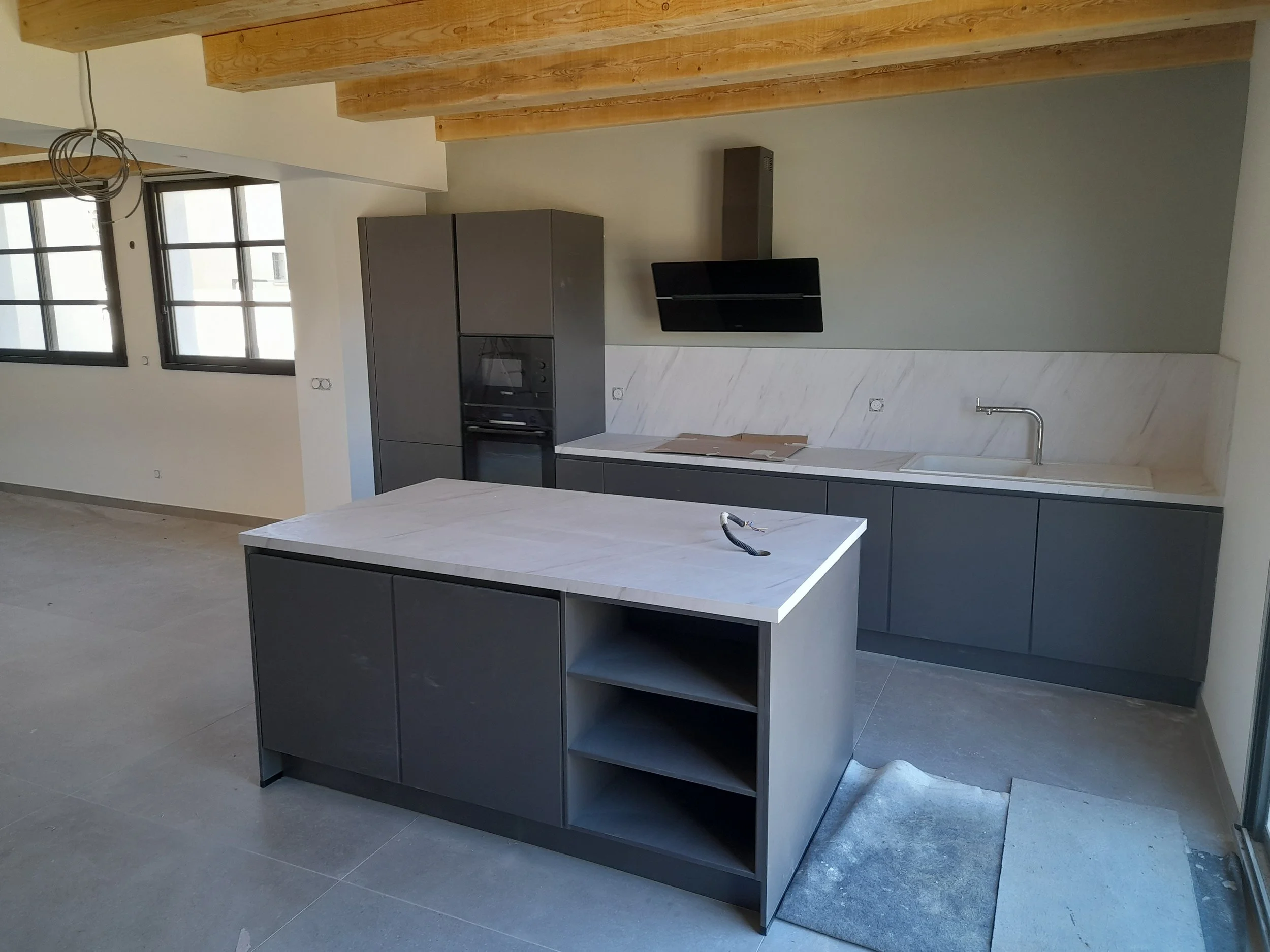 Cuisine moderne avec comptoir en marbre, armoires grises, fenêtres à cadre noir, plafond avec poutres en bois, et un évier en acier inoxydable.
