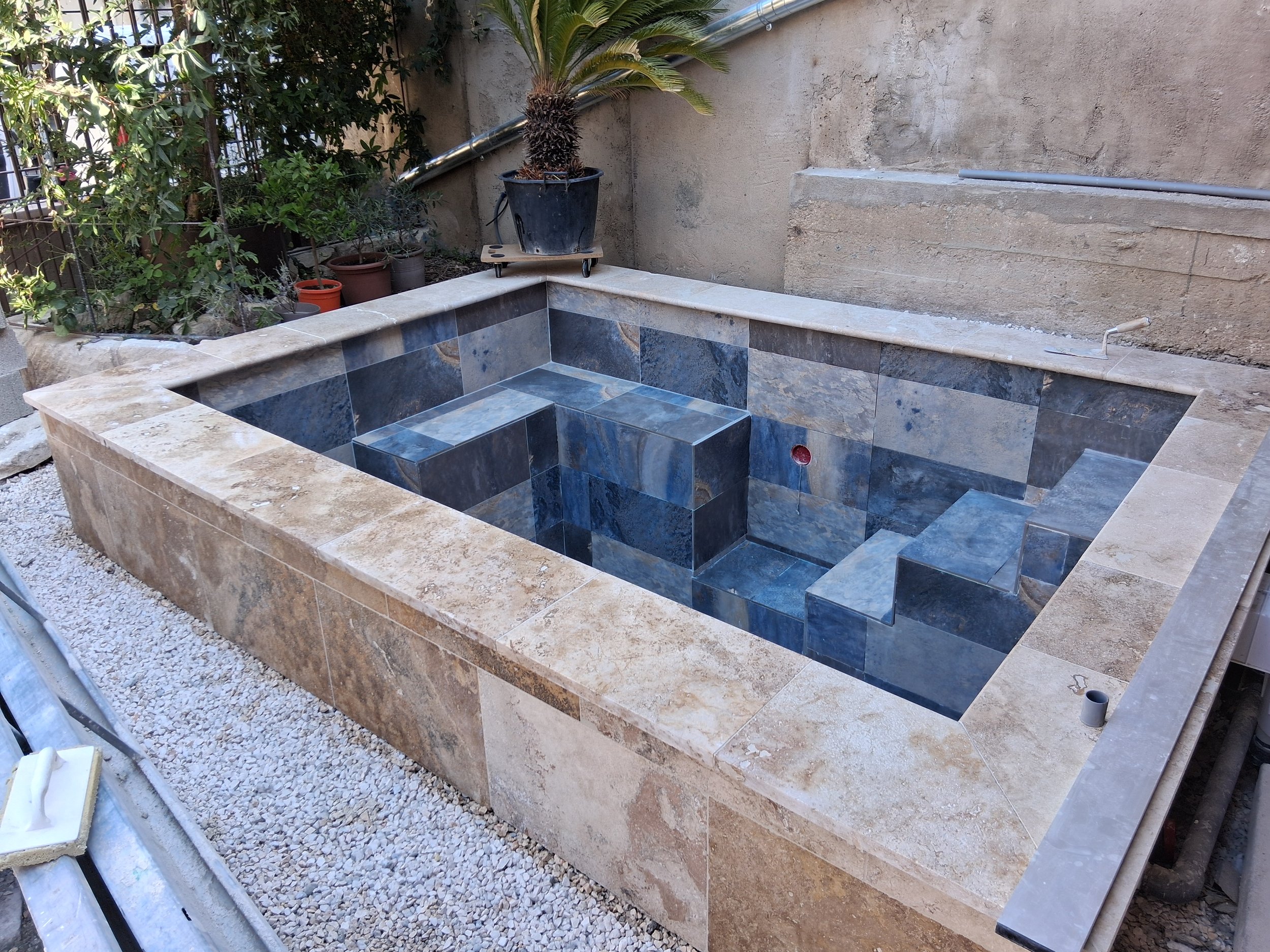 Une piscine en construction avec un revêtement en carreaux de pierre bleus et gris. Il y a des plantes vertes à l'arrière et des tuyaux de plomberie visibles sur le côté.
