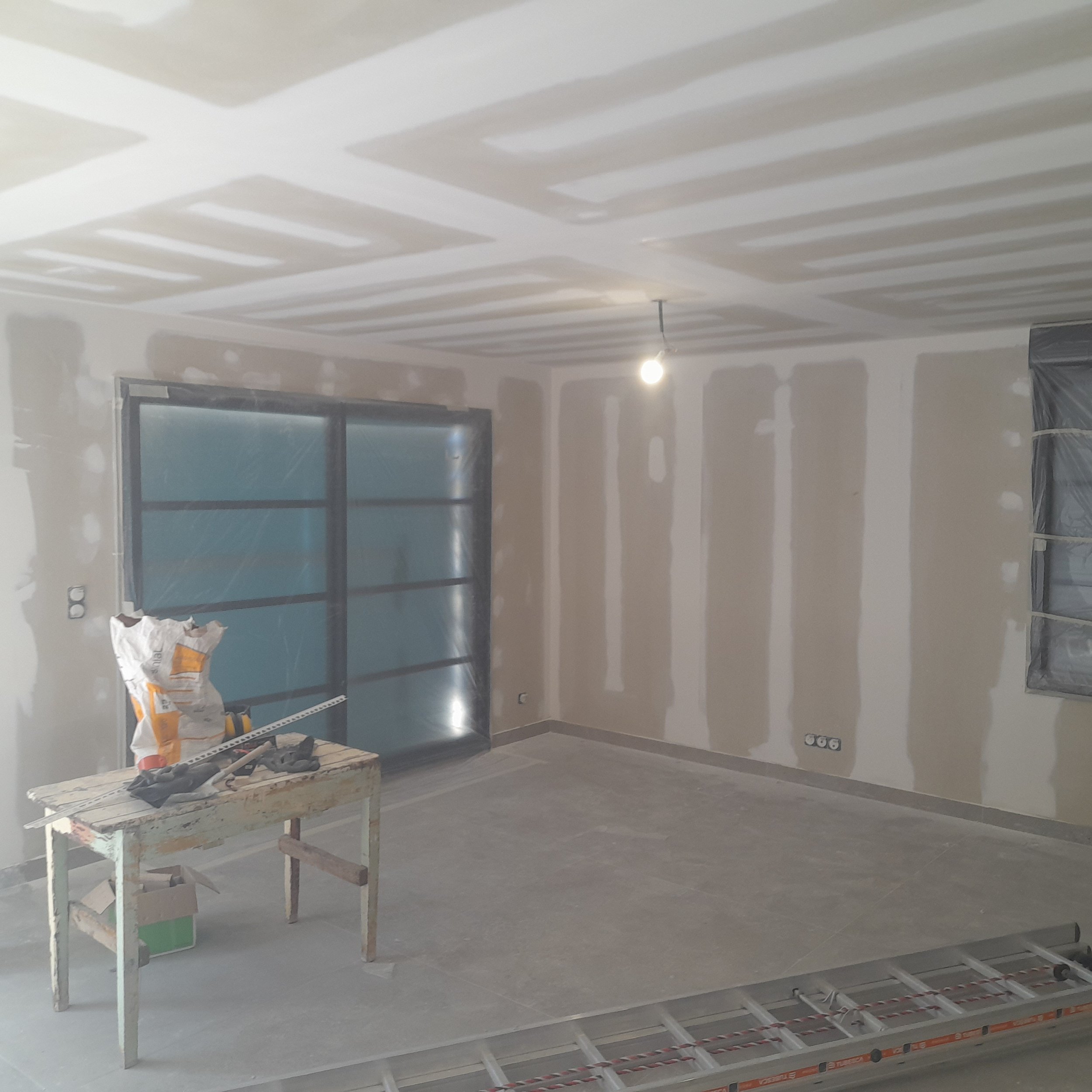 Une pièce en cours de rénovation avec des plafonds en cours de finition, un lampadaire allumé, une table en bois, des sacs de matériaux, et des murs en placoplâtre avec des prises électriques installées.