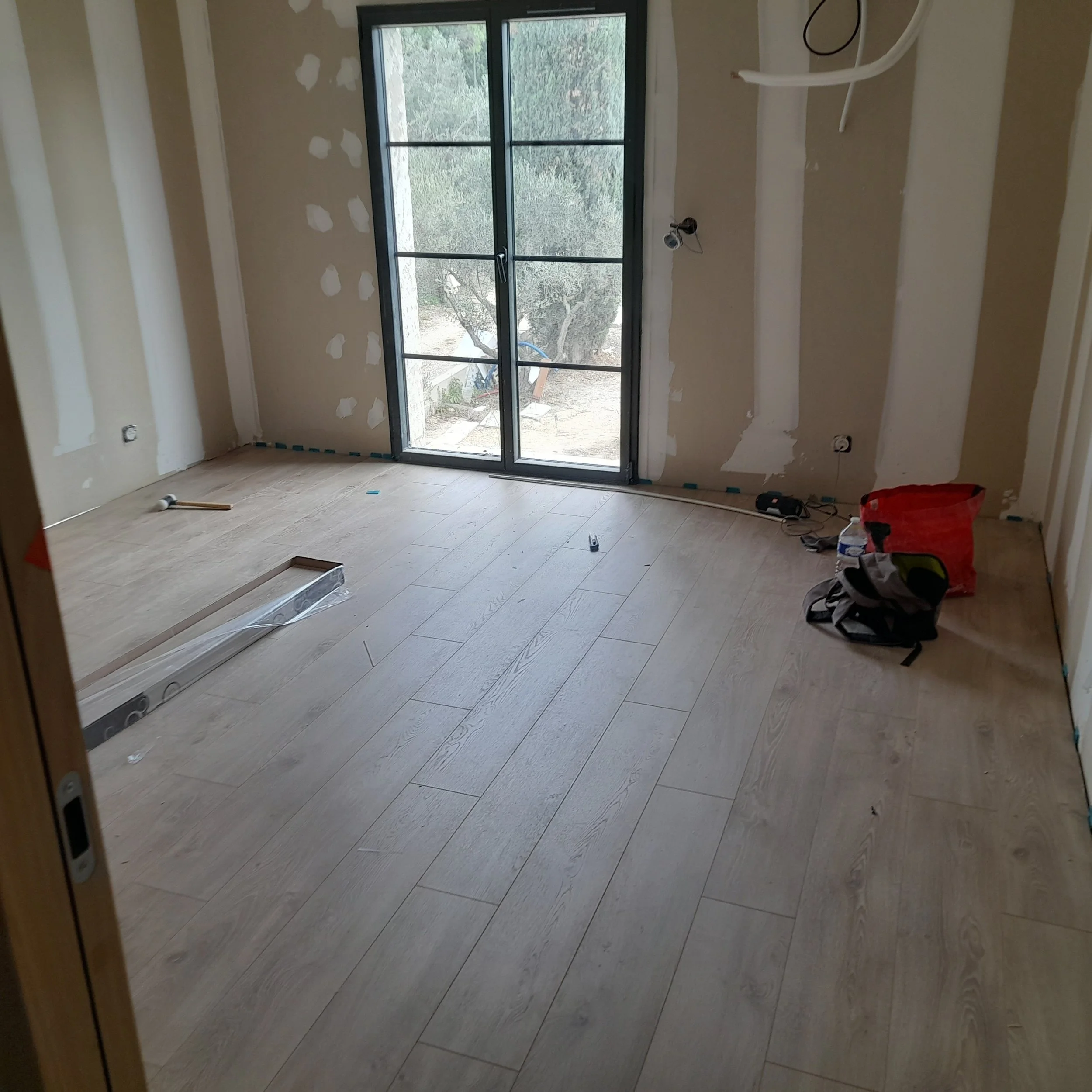 Intérieur d'une pièce en cours de rénovation avec un sol en bois clair, une porte-fenêtre en PVC noir donnant sur l'extérieur, des murs avec des plaques de plâtre en cours d'installation, et des outils de travail au sol.