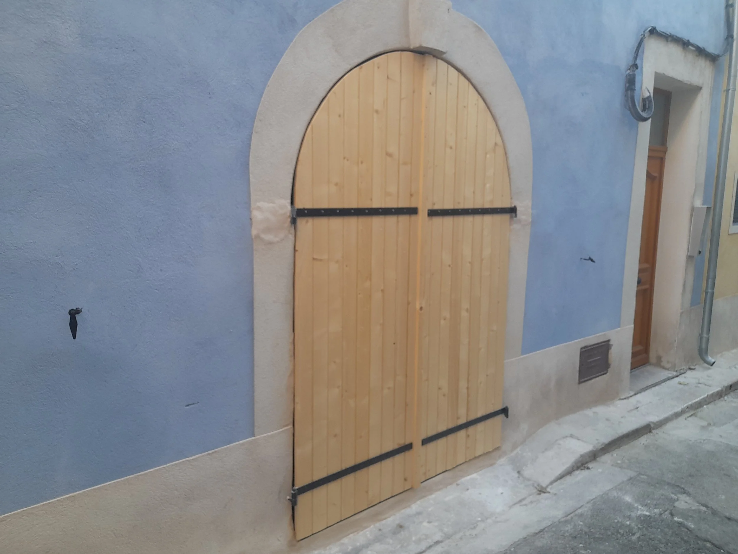 Une porte en bois en arc avec une bordure en béton, fixée à un mur bleu, avec une barre de verrouillage en bas dans un ancien bâtiment ou une maison en cours de rénovation.