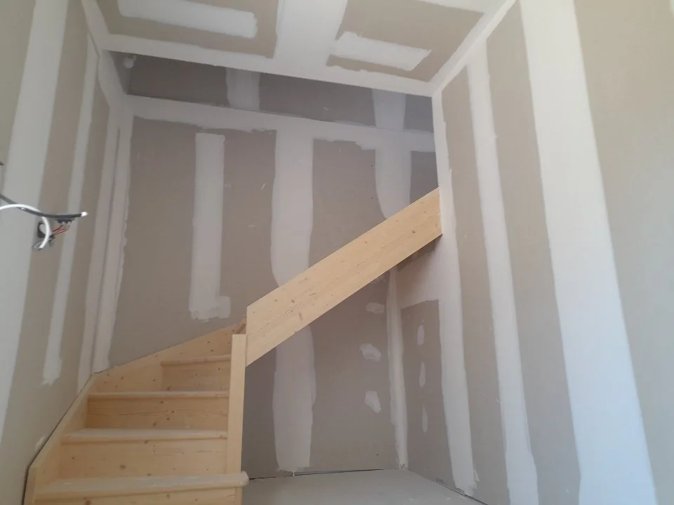 Un escalier en bois en construction dans une pièce avec murs en panneaux de gypse en cours de finition.