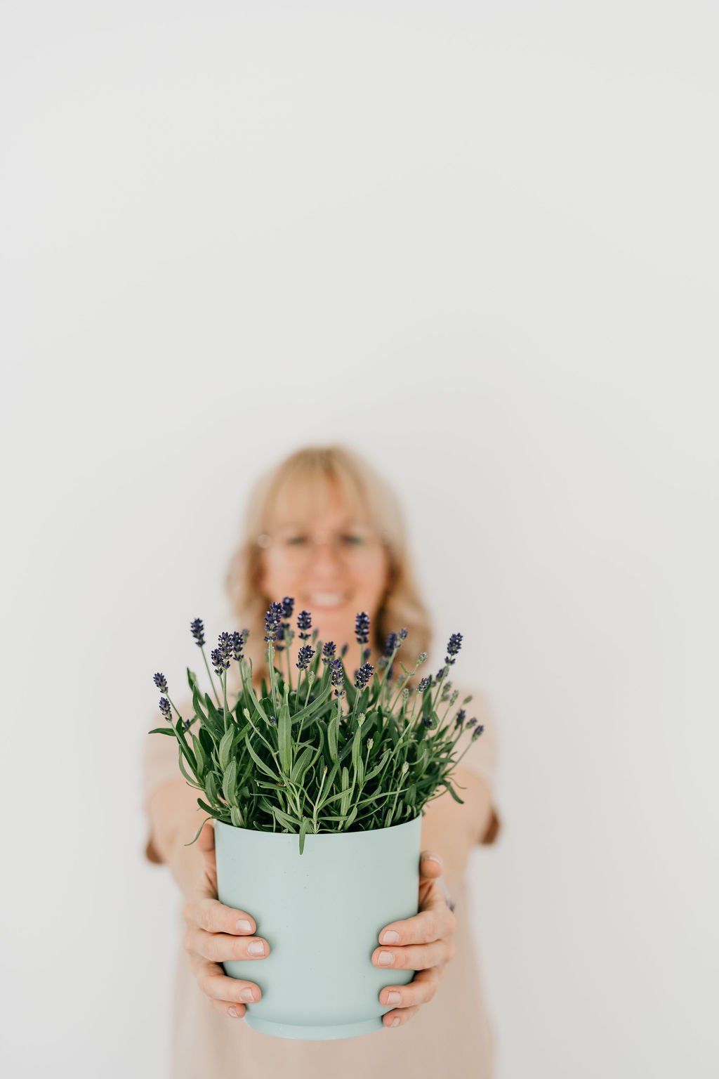 Frau hält eine Topfpflanze mit Lavendelblüten vor Kamera, Hintergrund ist weiß und unscharf.