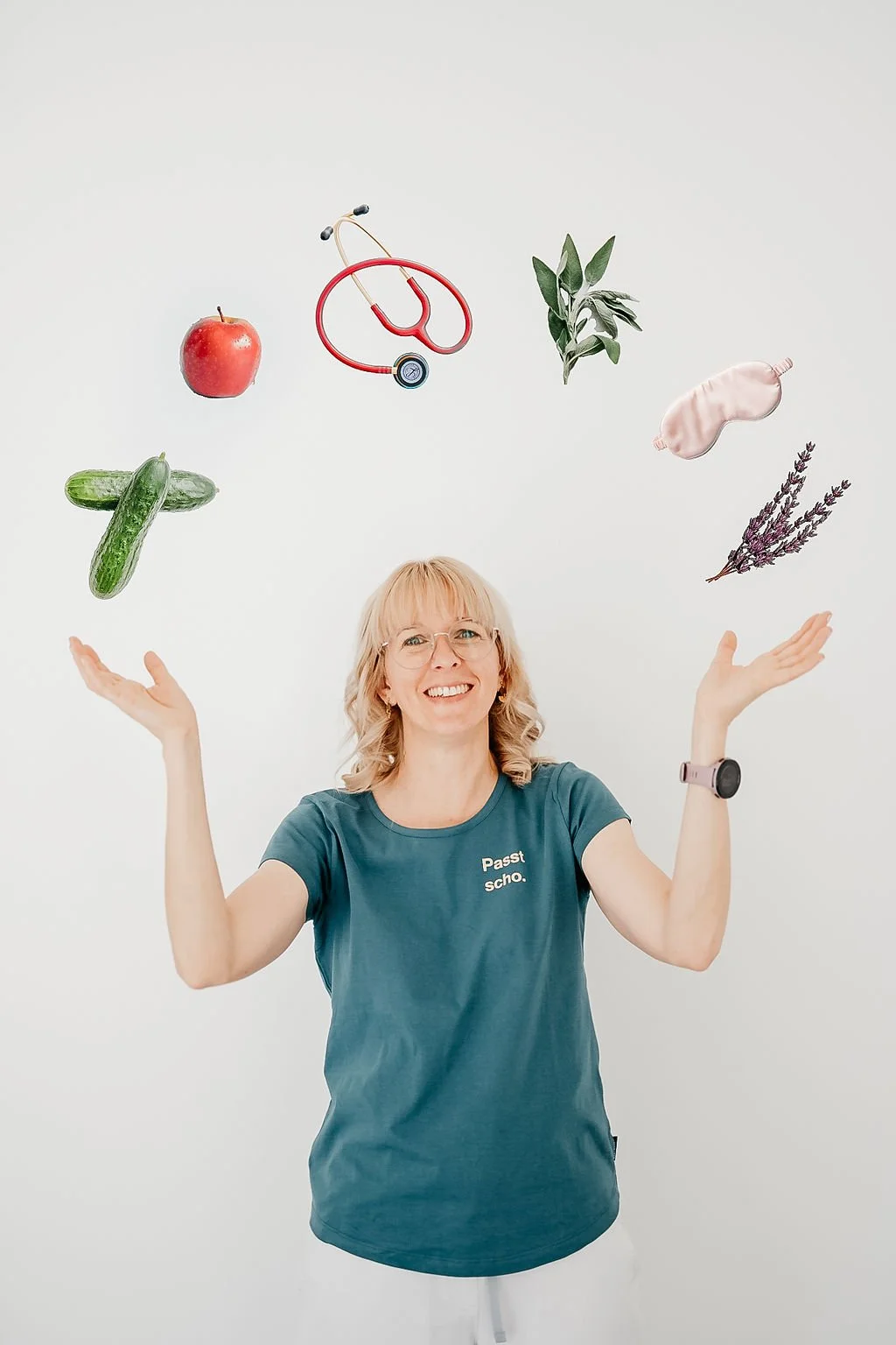 Eine lächelnde Frau trägt ein T-Shirt mit der Aufschrift 'Passt scho' und jongliert mit verschiedenen Gegenständen, darunter ein Apfel, eine Zucchini, eine Ohrhörer, Blätter, Lavendelsprühnebel, ein Stück Hühnchen und ein stethoskop.