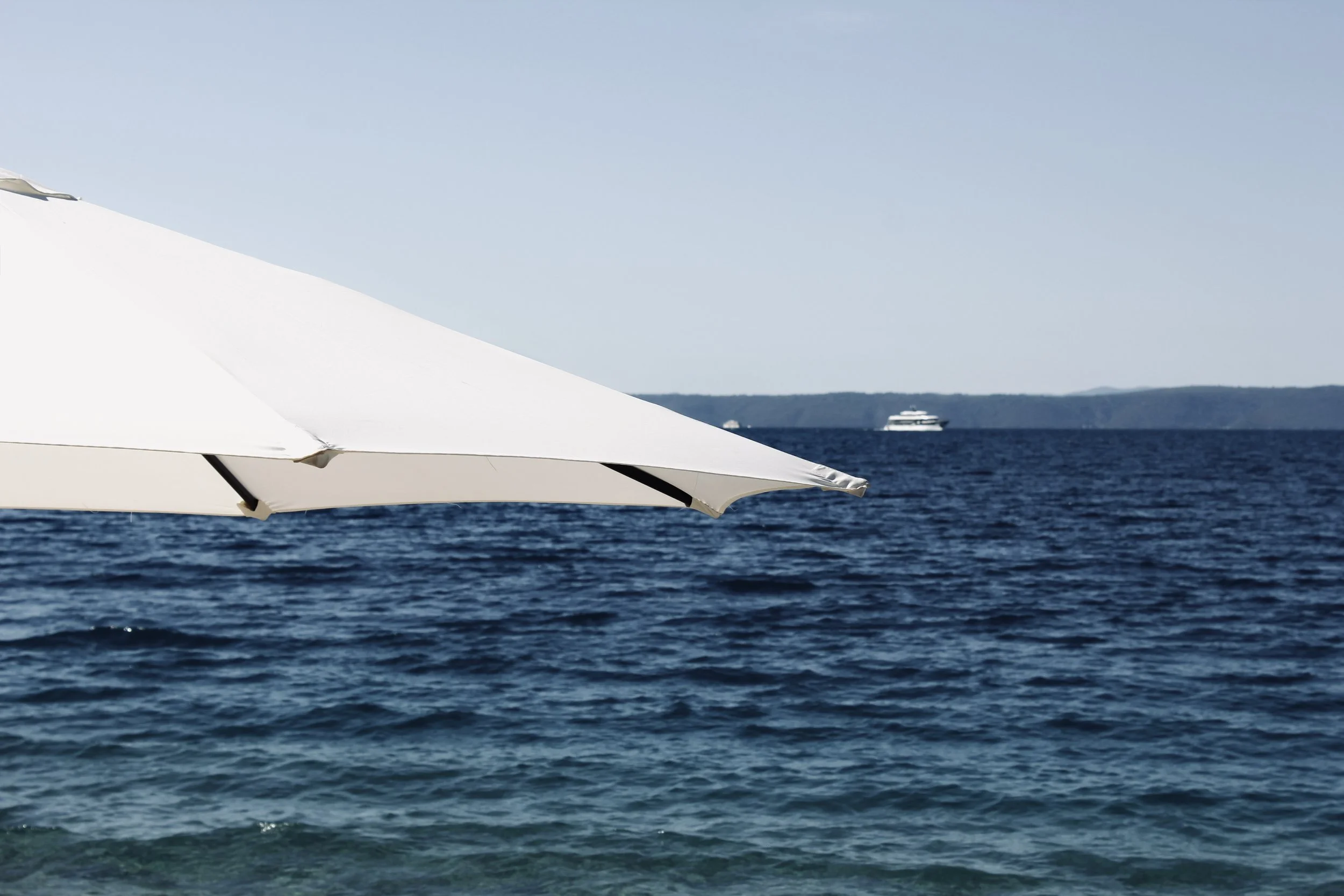 Part of a white outdoor umbrella over the ocean with a passenger boat in the distance on the water and hills on the horizon under a clear blue sky.