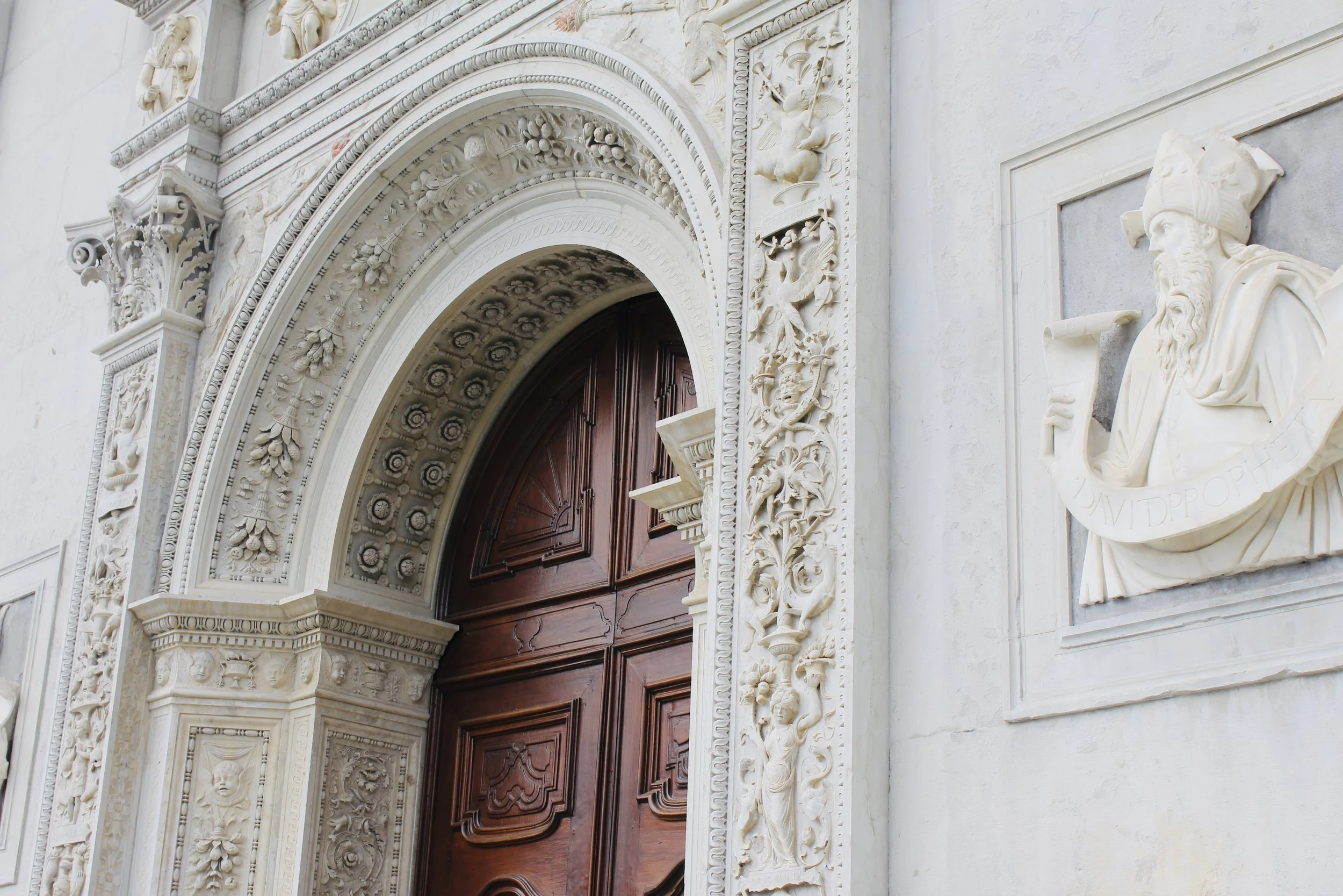 Close-up of ornate architectural detail around a grand wooden door, featuring intricate carved patterns, floral motifs, and a sculpture of a bearded man in a classical style.