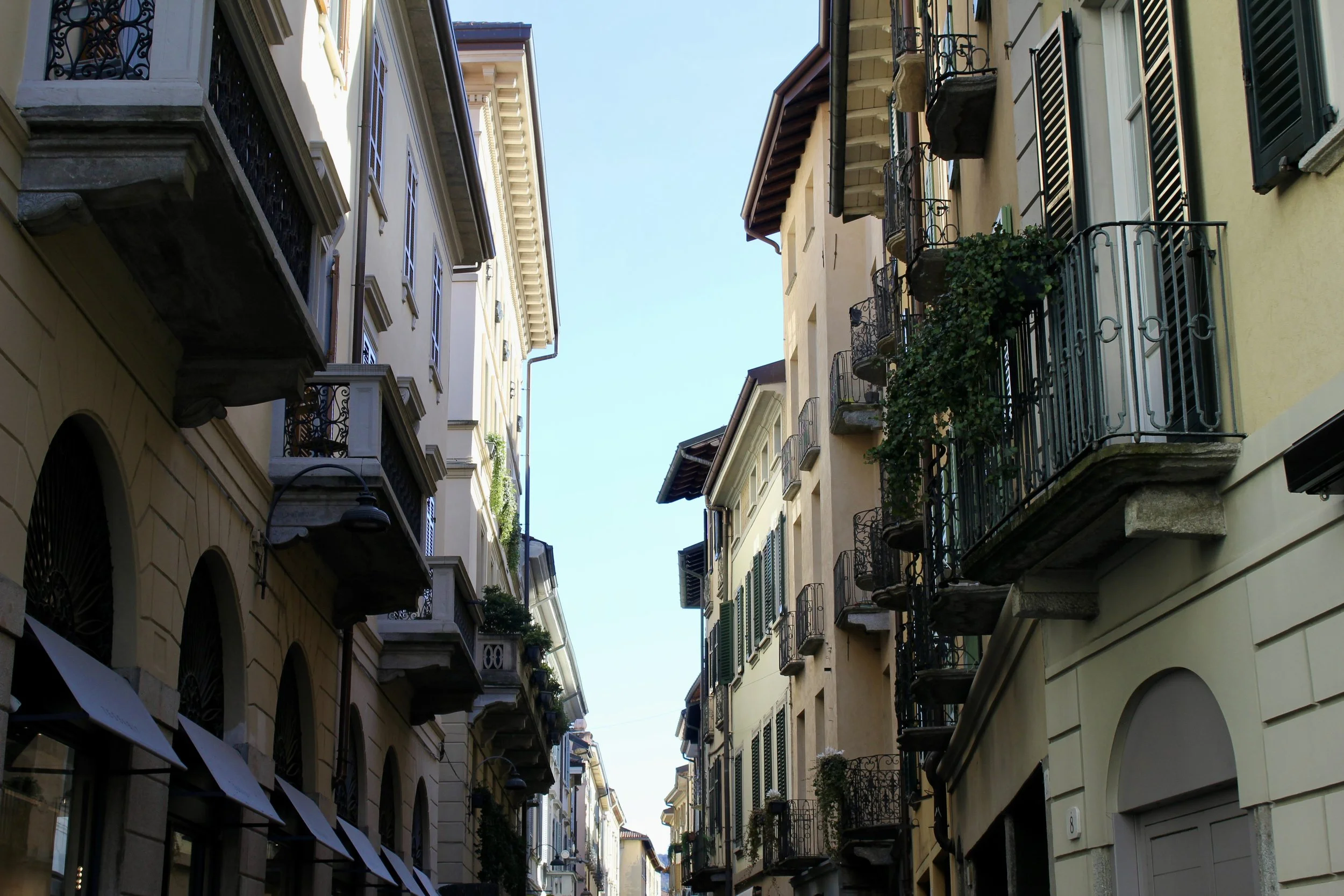 A narrow street in a European city with multi-story buildings on either side, featuring balconies with wrought iron railings, green shutters, and some with plants. The sky is clear with a few clouds.