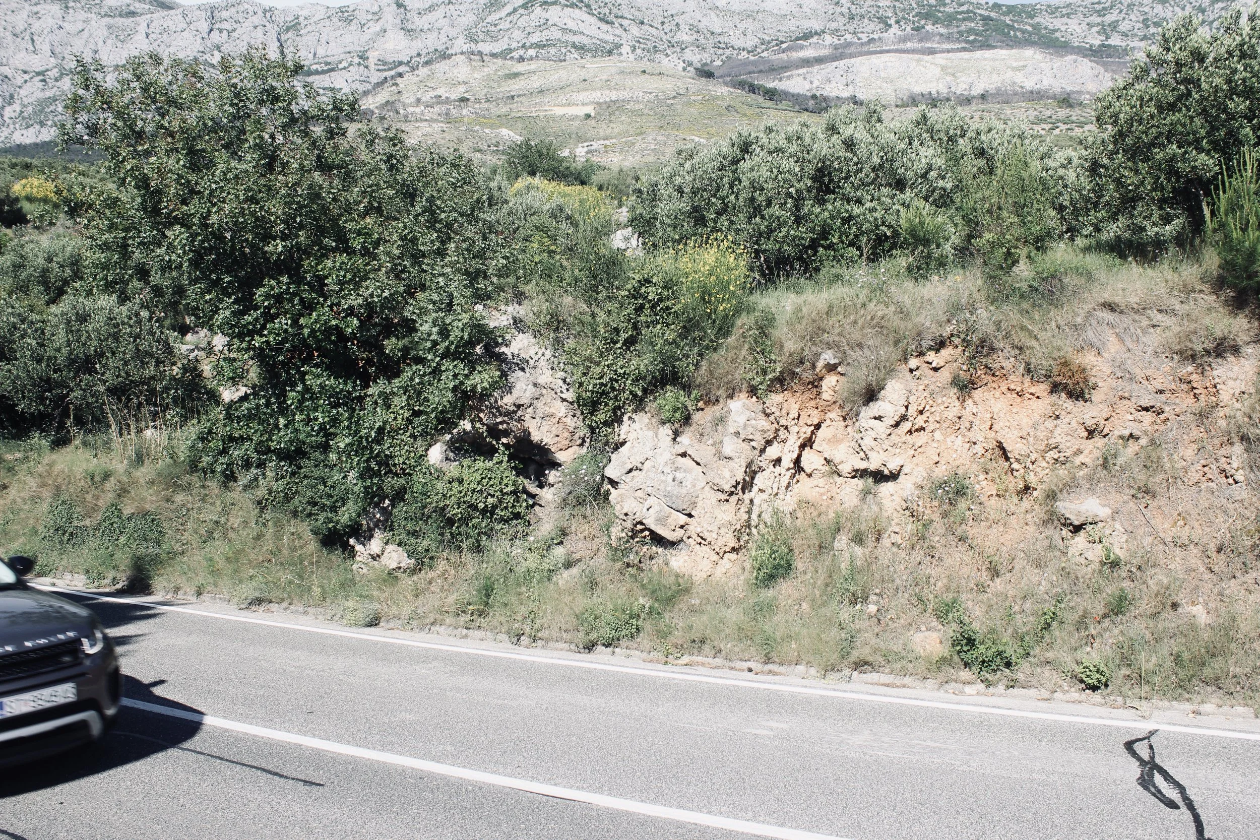 A mountain landscape with green shrubbery and rocky terrain viewed from a road, with a part of a black car visible in the lower left corner.