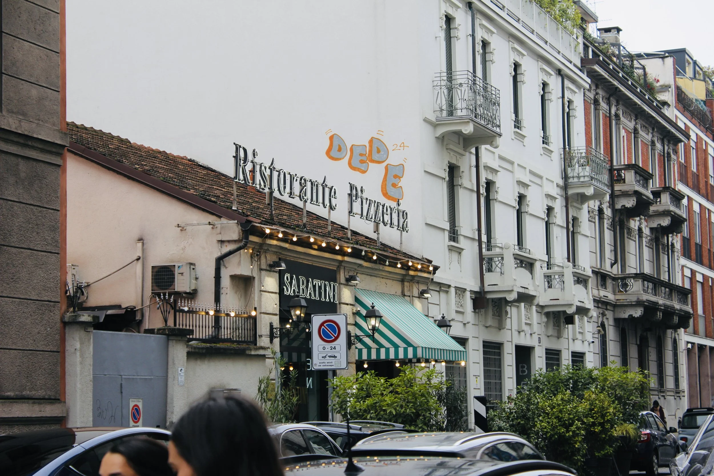 Street view showing a restaurant named Rickstee Pizzeria with outdoor seating, parked cars, and multi-story buildings with balconies in the background.