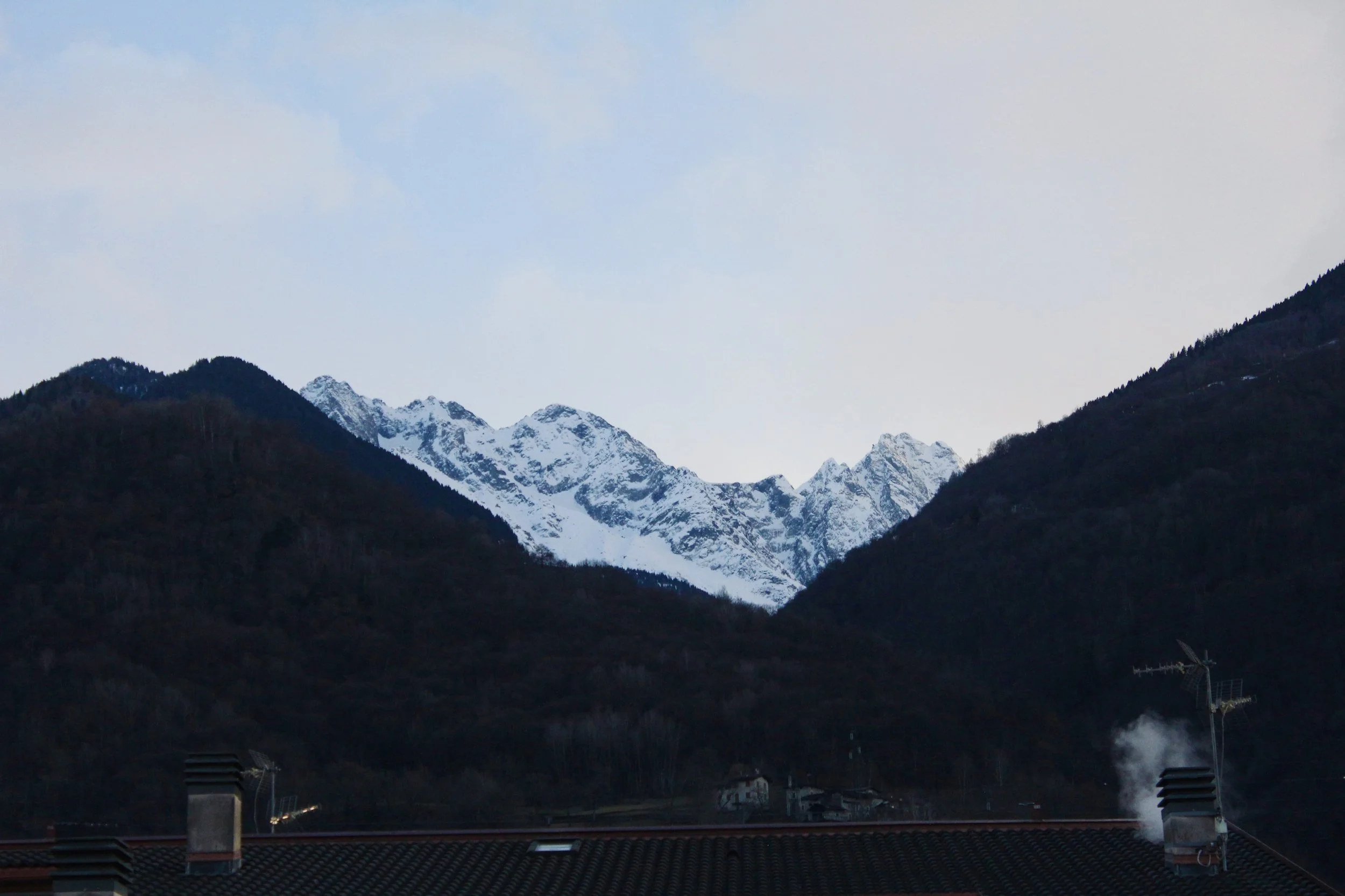 Mountain range with snow-capped peaks, viewed from a small town or village with rooftops and chimneys in the foreground.