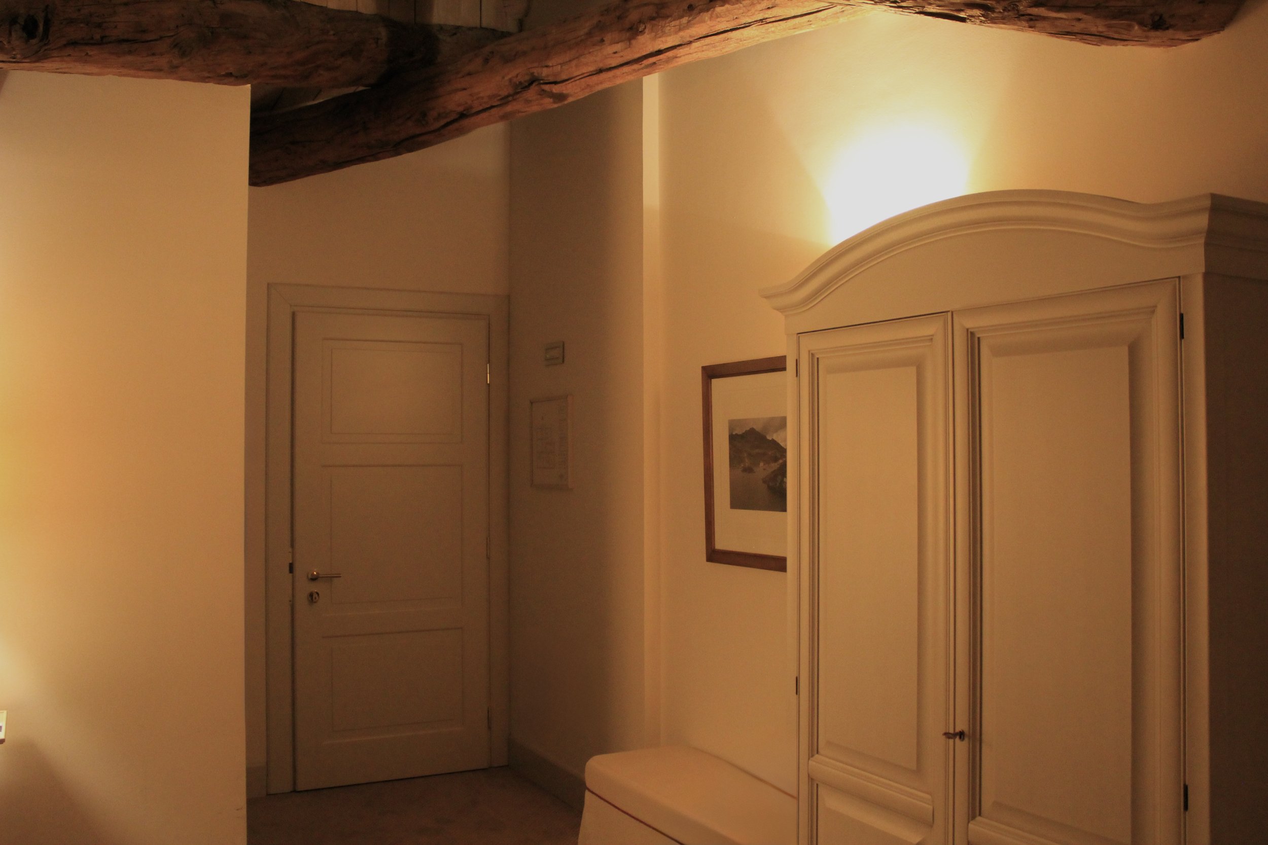 Interior of a room with beige walls, a white armoire, framed artwork on the wall, wooden beams on the ceiling, and a door on the left side.