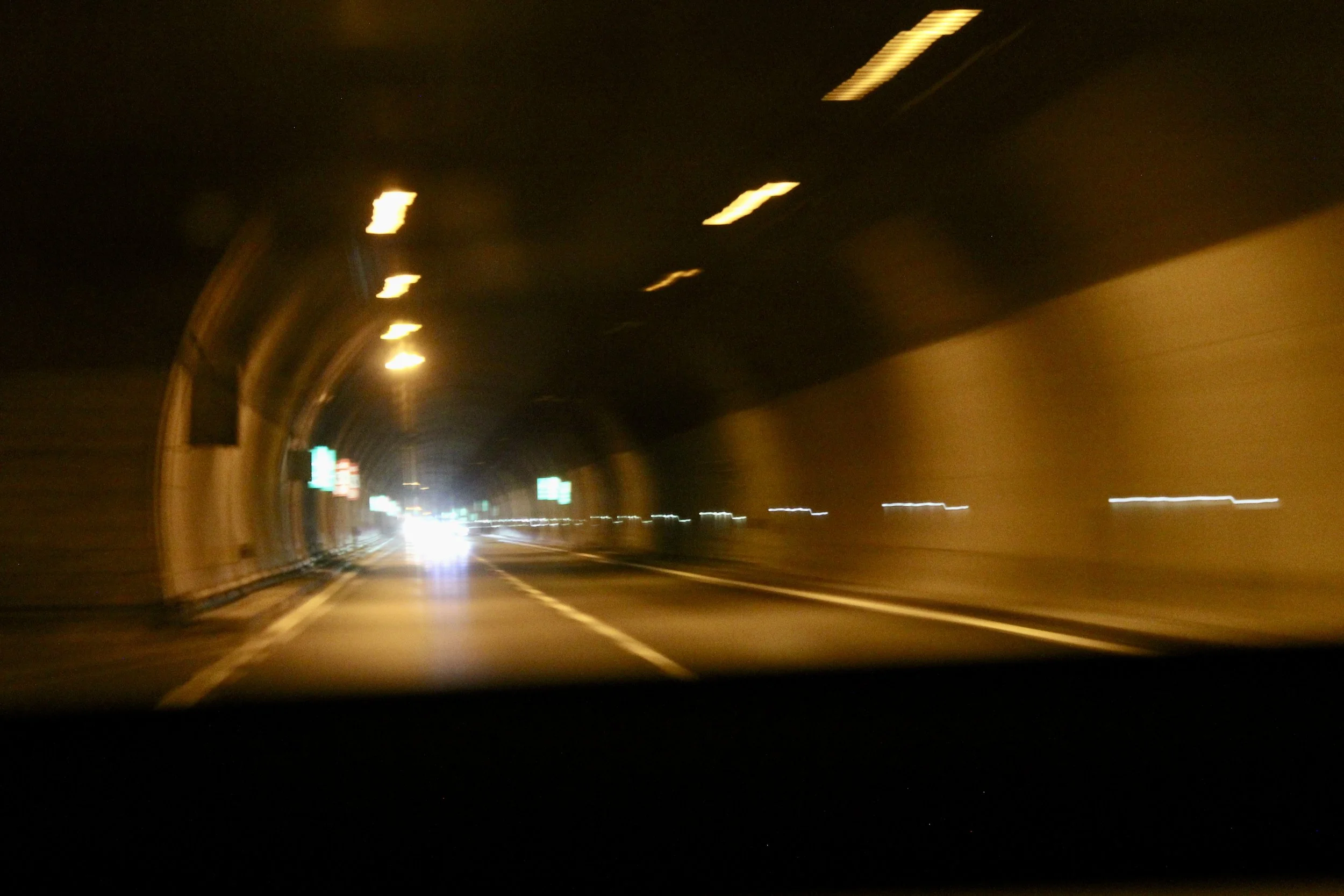 Photo of a dark tunnel or passageway with yellow lighting and a curving road, with some blurred lights in the distance, taken from inside a moving vehicle.