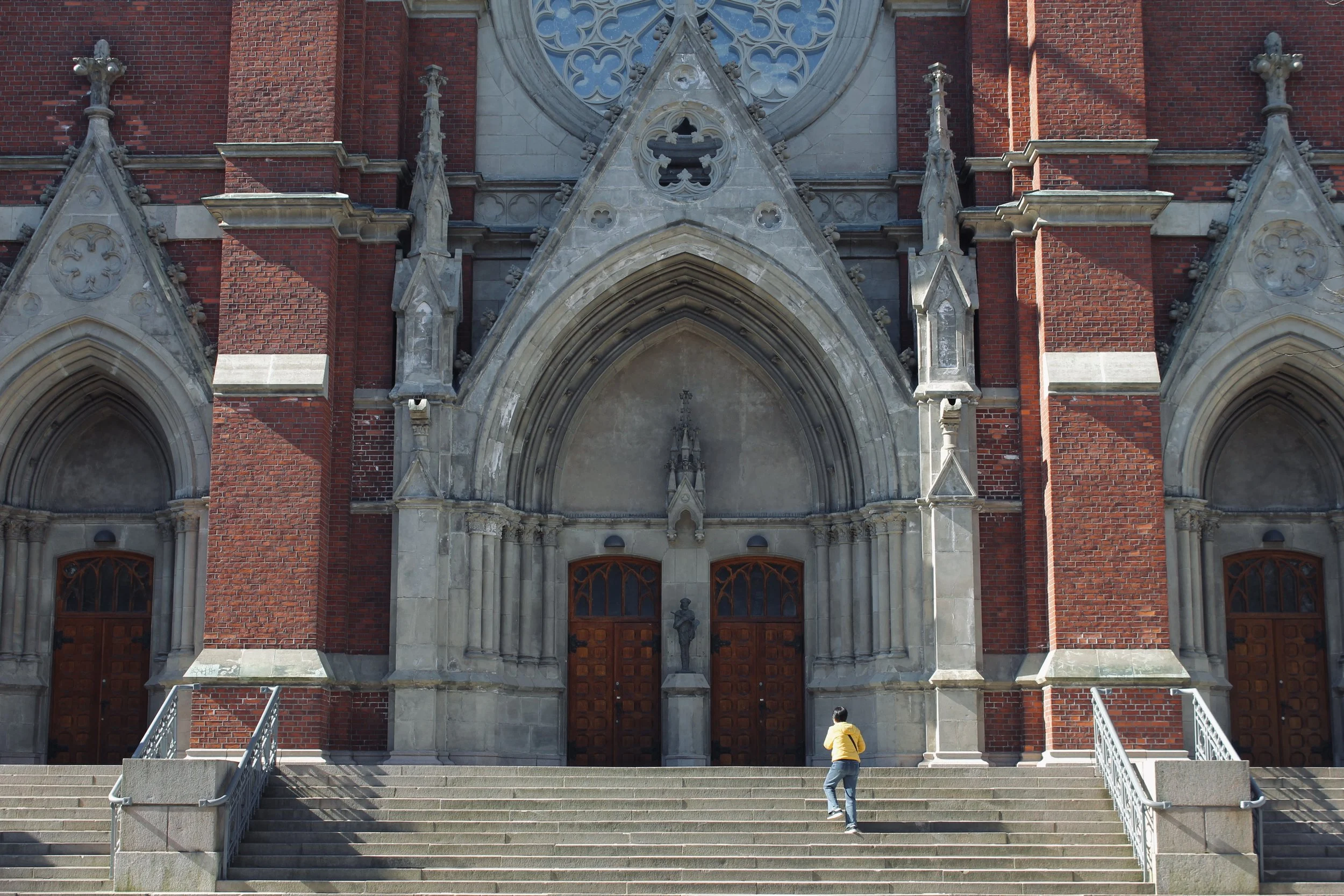 Front view of a Gothic-style church with red brick walls, stone arches, and stained glass windows, with two people on the stairs leading to the entrance.