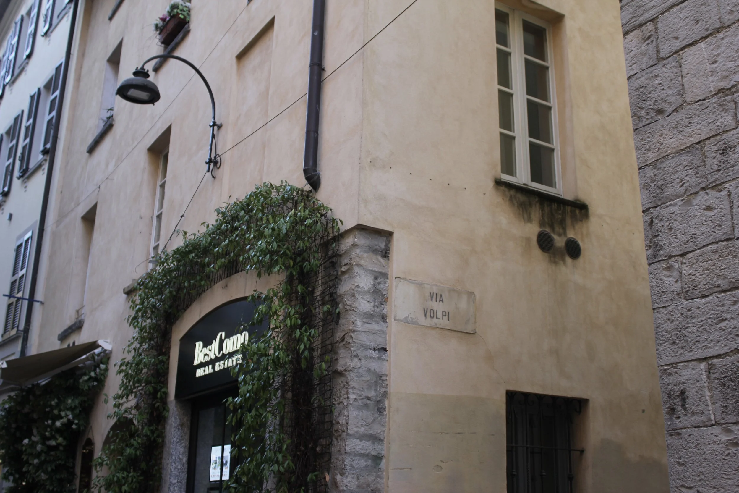 A narrow street corner with beige and gray stone buildings, a street sign that reads 'VIA VOLPI,' a black street lamp, and a sign for a restaurant called 'BastCome' with green foliage and flowers above the entrance.