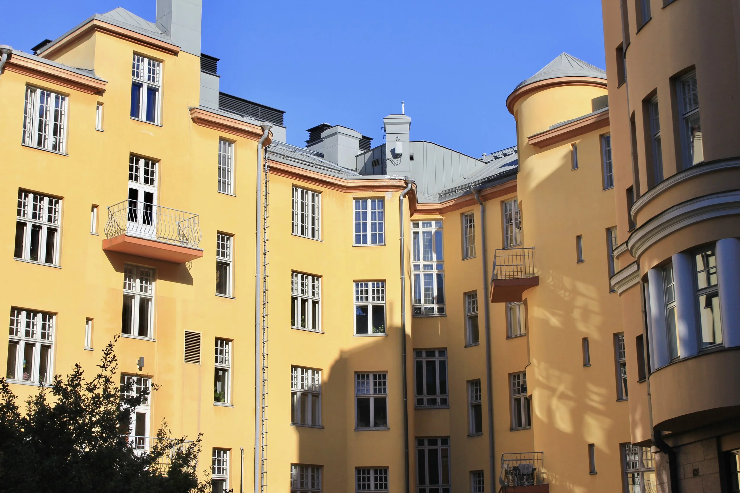 This image depicts a brightly colored residential building, primarily painted in yellow with several windows and small balconies. The roof is metallic gray with multiple chimneys and vents. The clear blue sky serves as a backdrop, and some plants are
