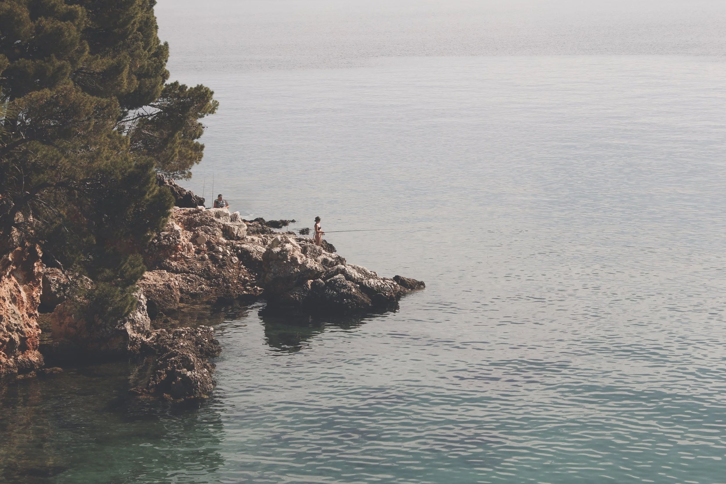 People fishing off rocky shoreline with trees on the left and water extending to the horizon.