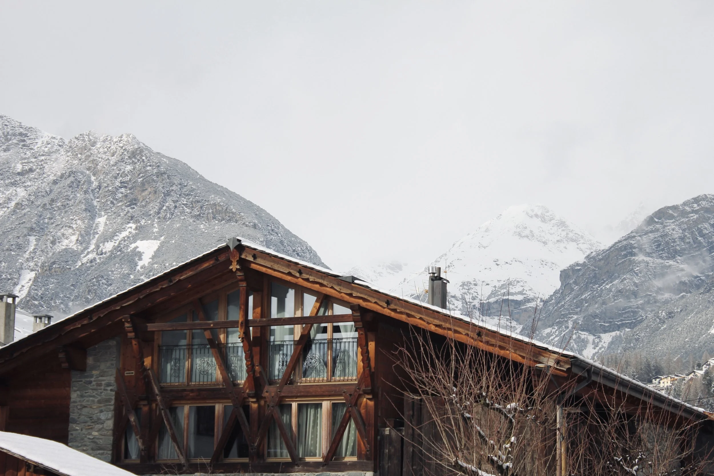 A mountain chalet with wooden and stone architecture set against snow-covered mountains and a cloudy sky.