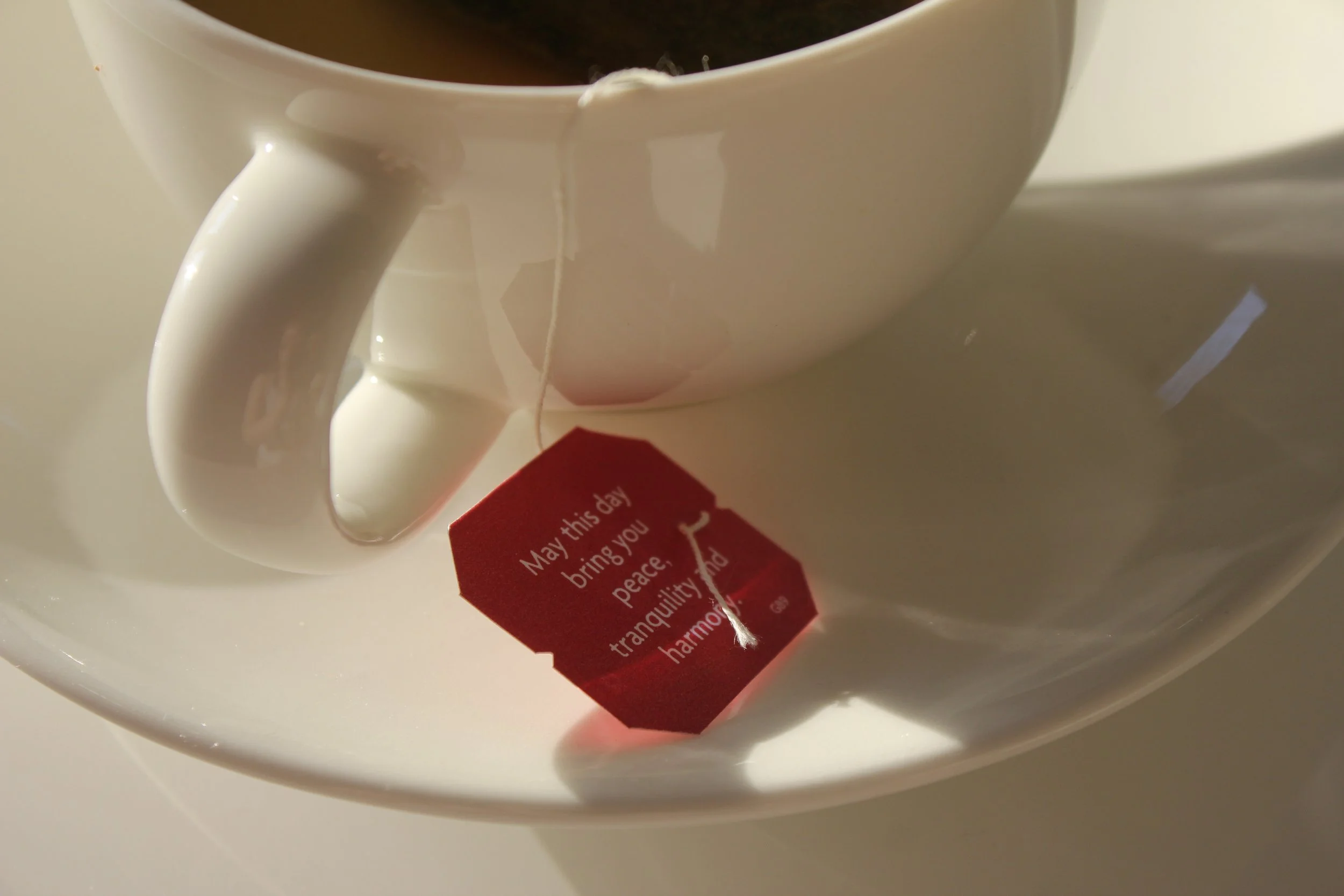 Close-up of a white ceramic mug filled with dark tea, with a red tag attached that reads "May this day bring you peace, tranquility, and harmony." The mug has a smooth, curved handle and is placed on a white surface.