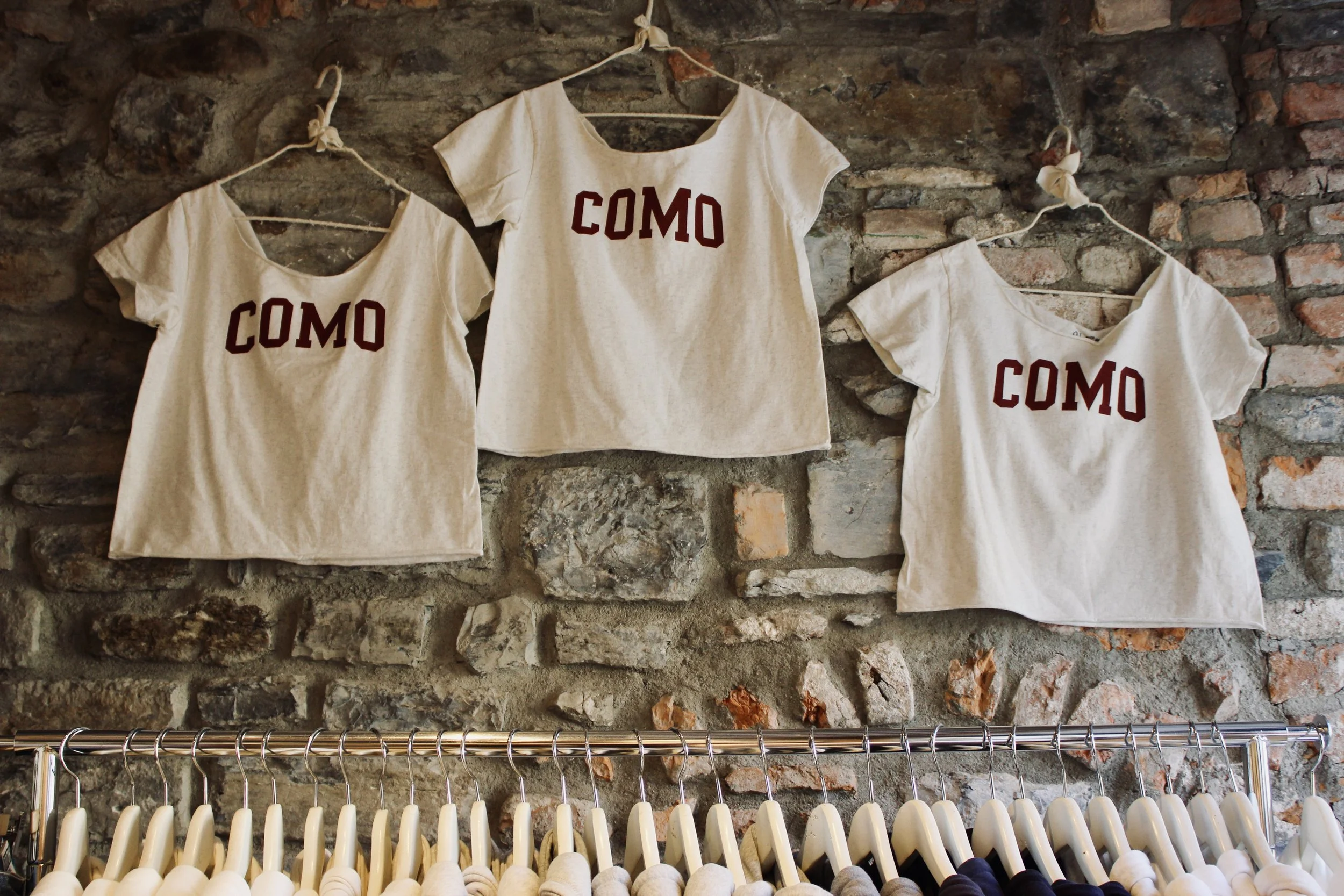 Three beige T-shirts with the word 'COMO' printed in red, hanging on a rustic brick wall. The shirts are on hangers connected with a string, with a row of white and beige clothing hangers below near a metal rack.