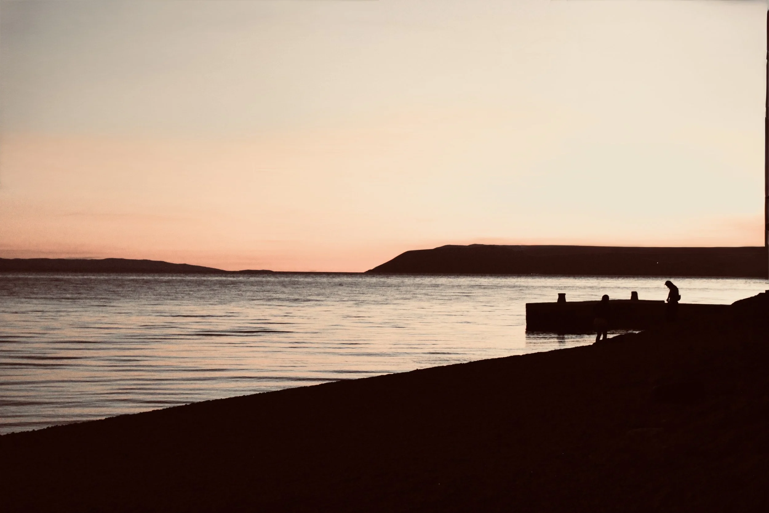 A sunset view over a calm body of water with a cliff on the horizon, silhouetted by the setting sun. A person stands on a pier near the shore, looking at the water.